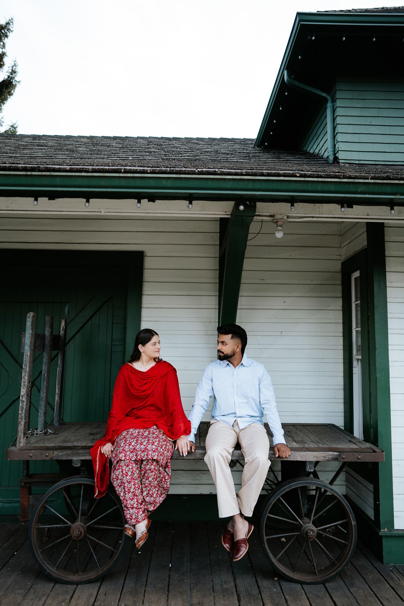 Couple sitting on rustic wooden cart on porch of historic building, woman in red outfit, man in light blue shirt.
