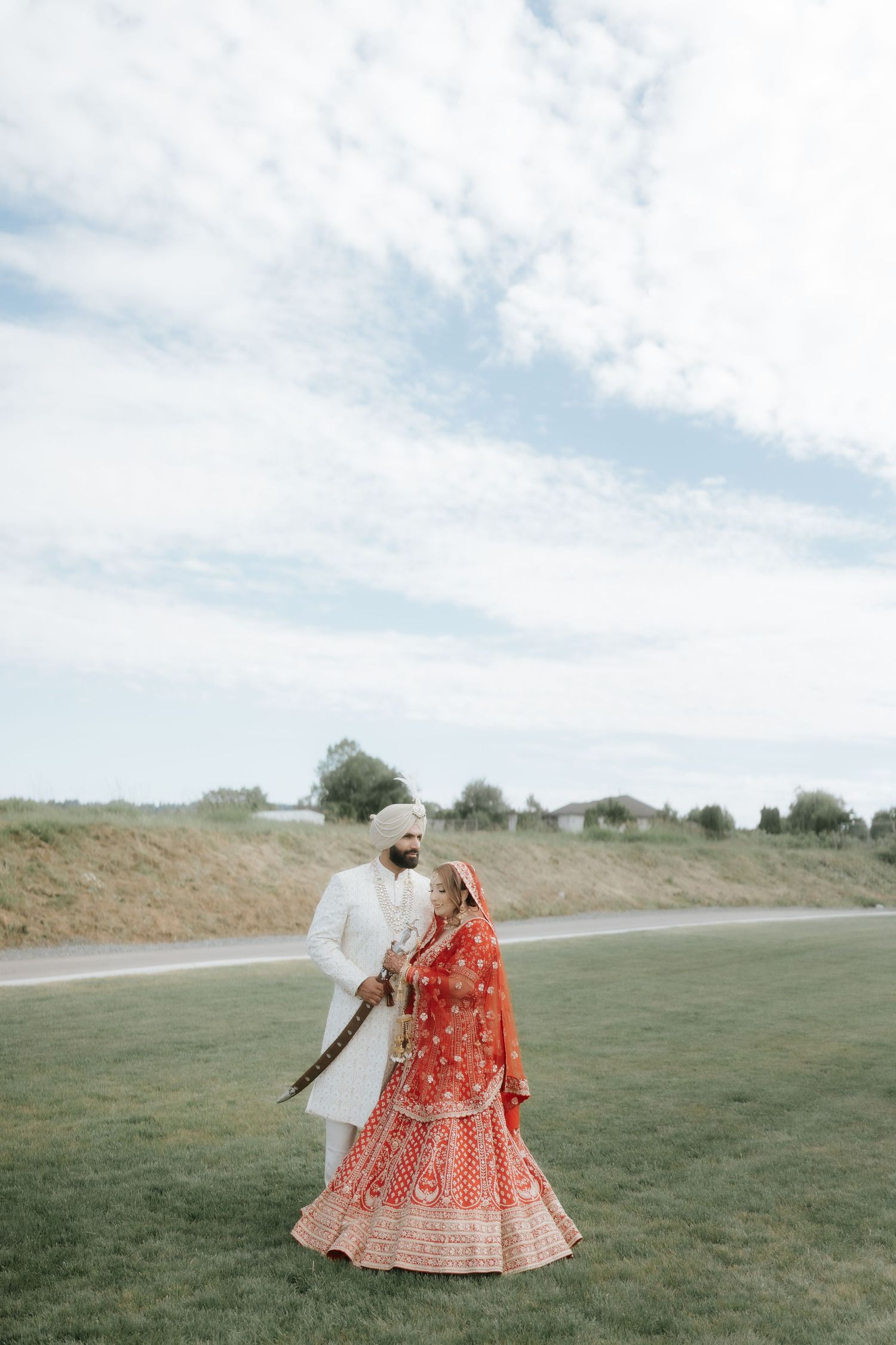 A Sikh bride in red lehenga and groom in white sherwani share a romantic moment outdoors on a cloudy day.