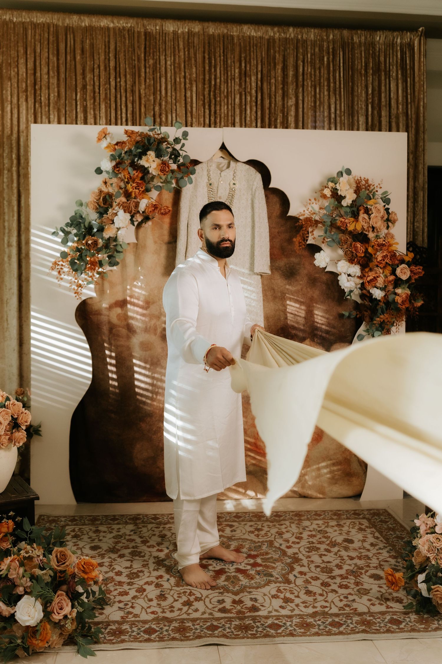A bearded groom in white kurta stands before a floral backdrop with his sherwani displayed at an Indian wedding.
