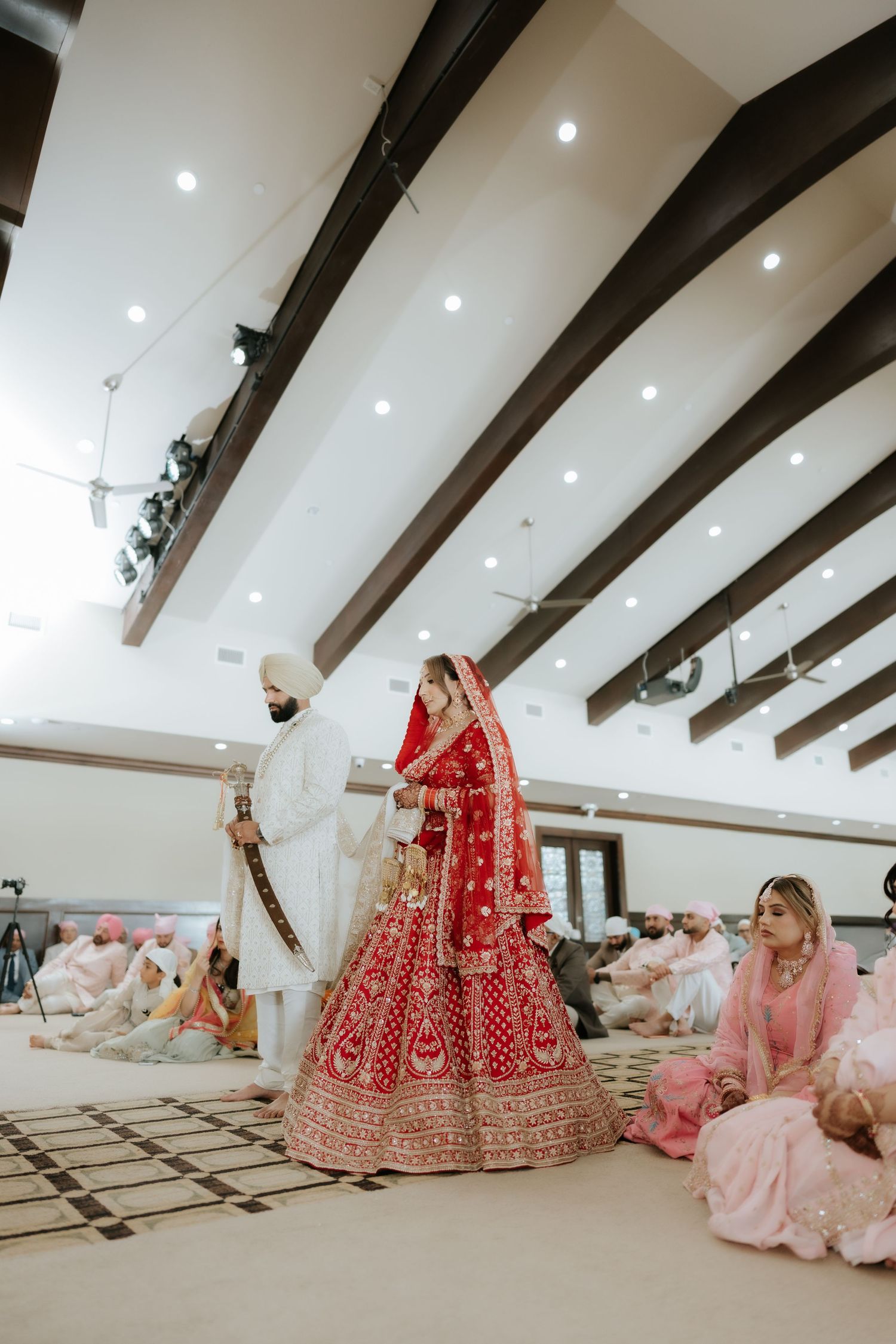 Sikh bride in red lehenga and groom in white sherwani performing Anand Karaj wedding ceremony in a gurudwara.