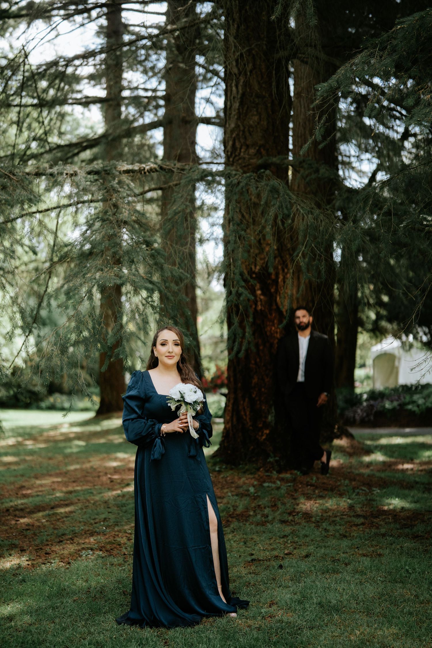 Woman in teal gown holds bouquet in forest, man in dark suit stands behind large tree in romantic outdoor setting.