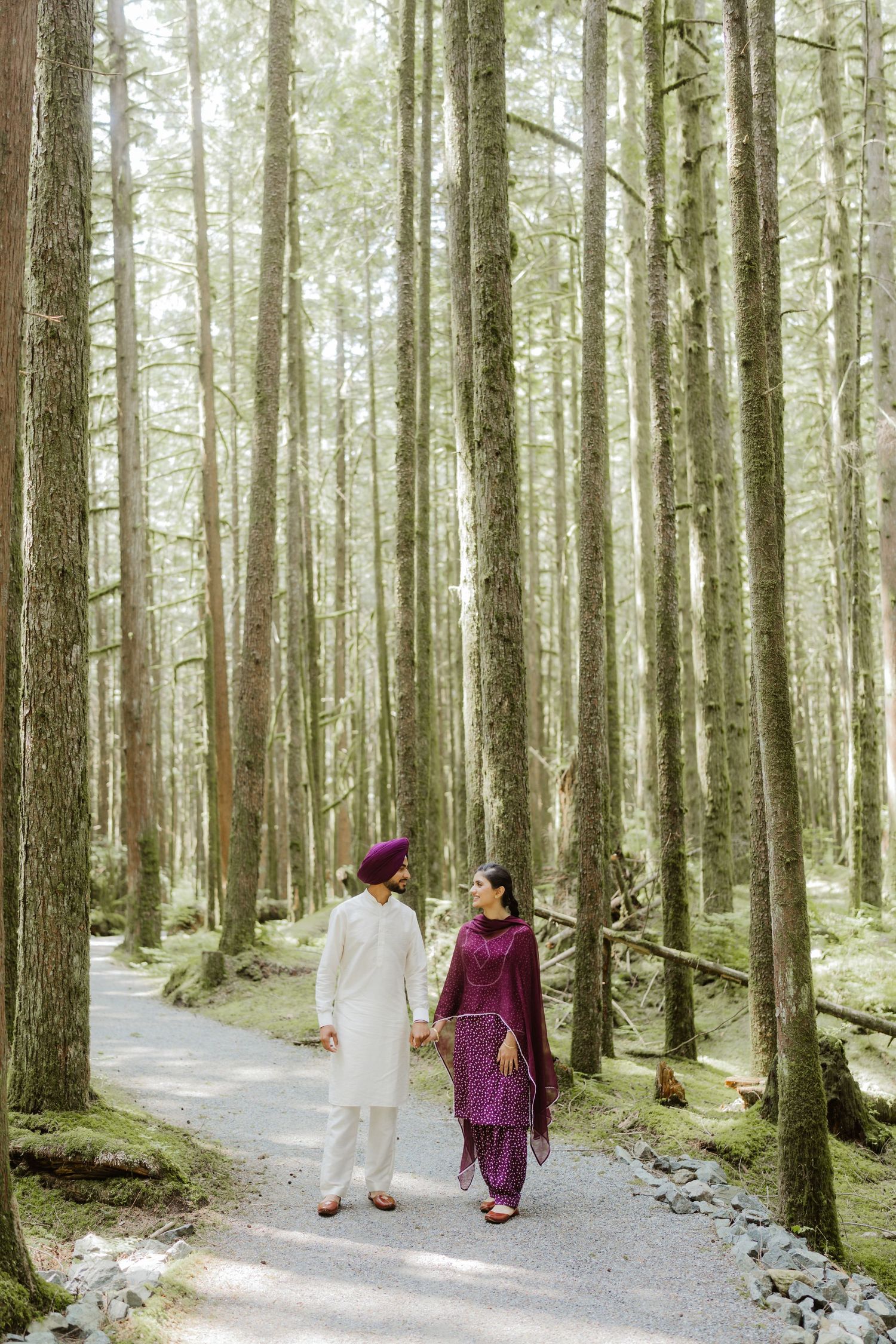 Sikh couple in traditional attire walking hand-in-hand along a forest path surrounded by tall trees.