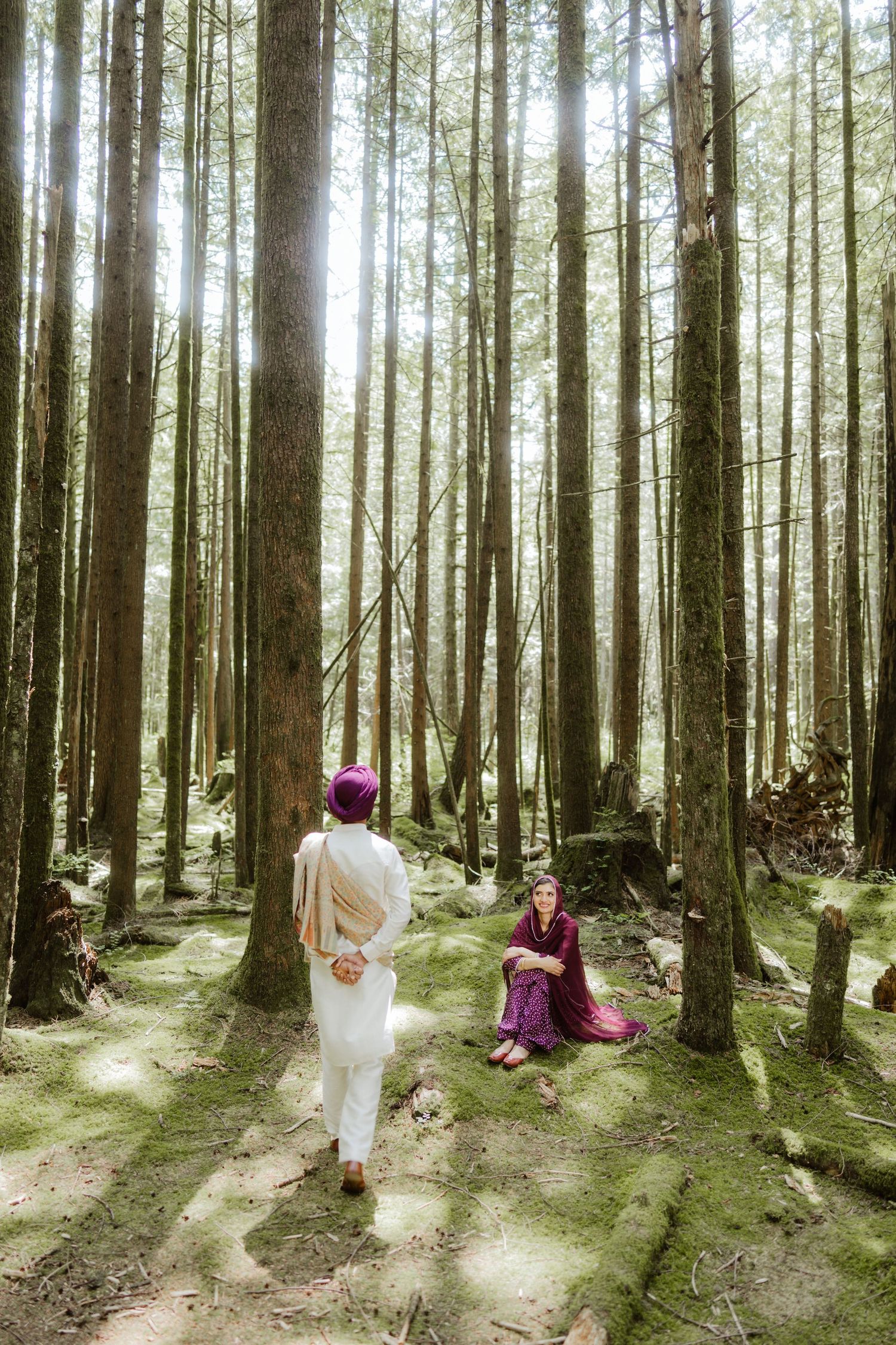 Sikh man approaching woman sitting on mossy ground in a sunlit forest with tall trees.
