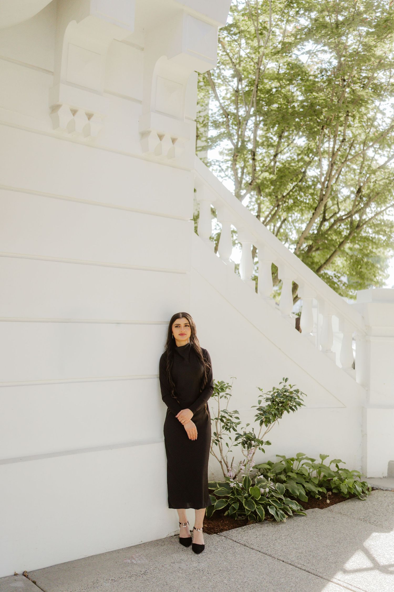 Woman in black long-sleeve midi dress leaning against a white balustrade wall outdoors.