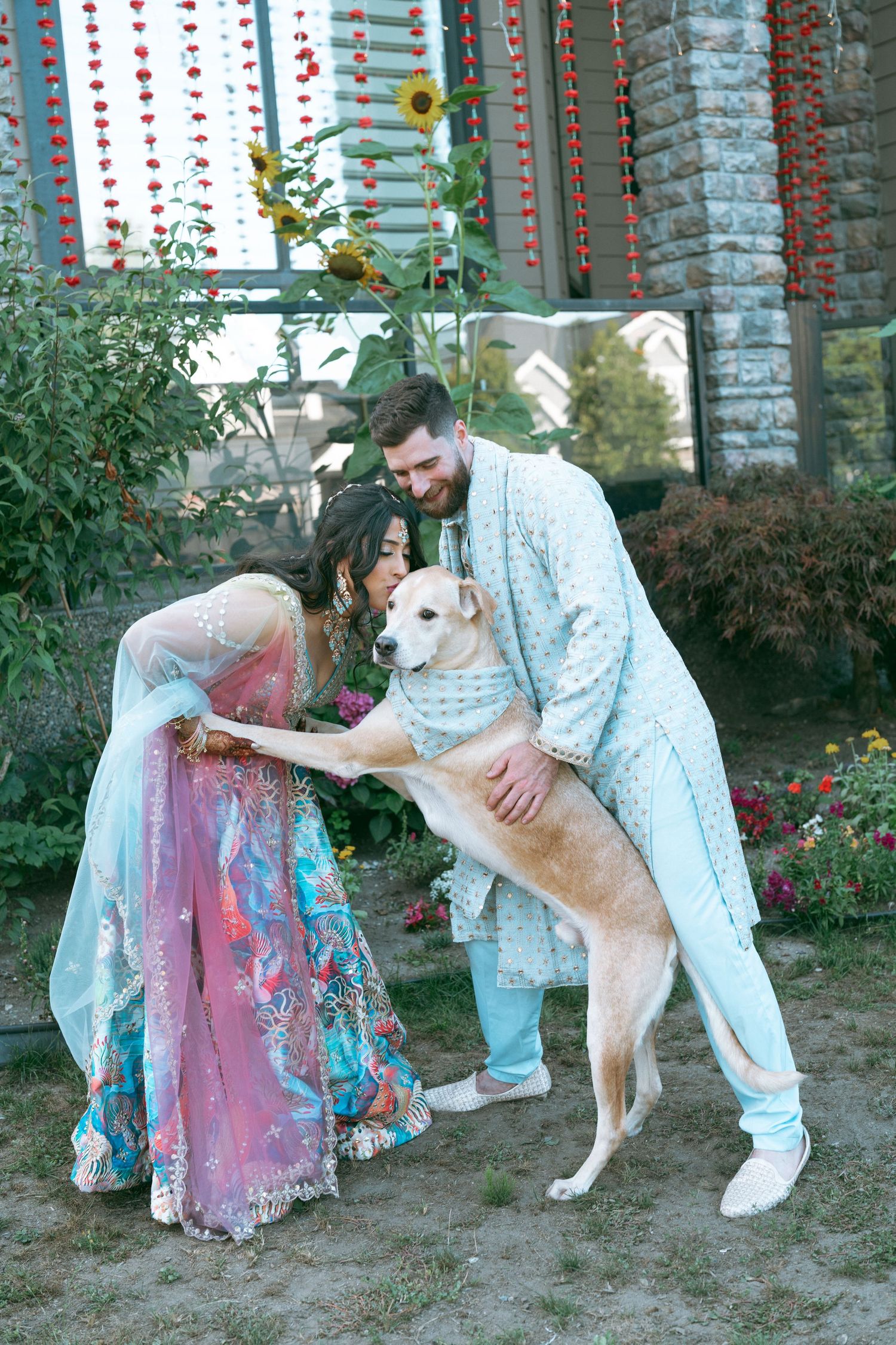 Couple in traditional South Asian wedding attire playfully interacting with a dog outdoors, surrounded by floral decorations.