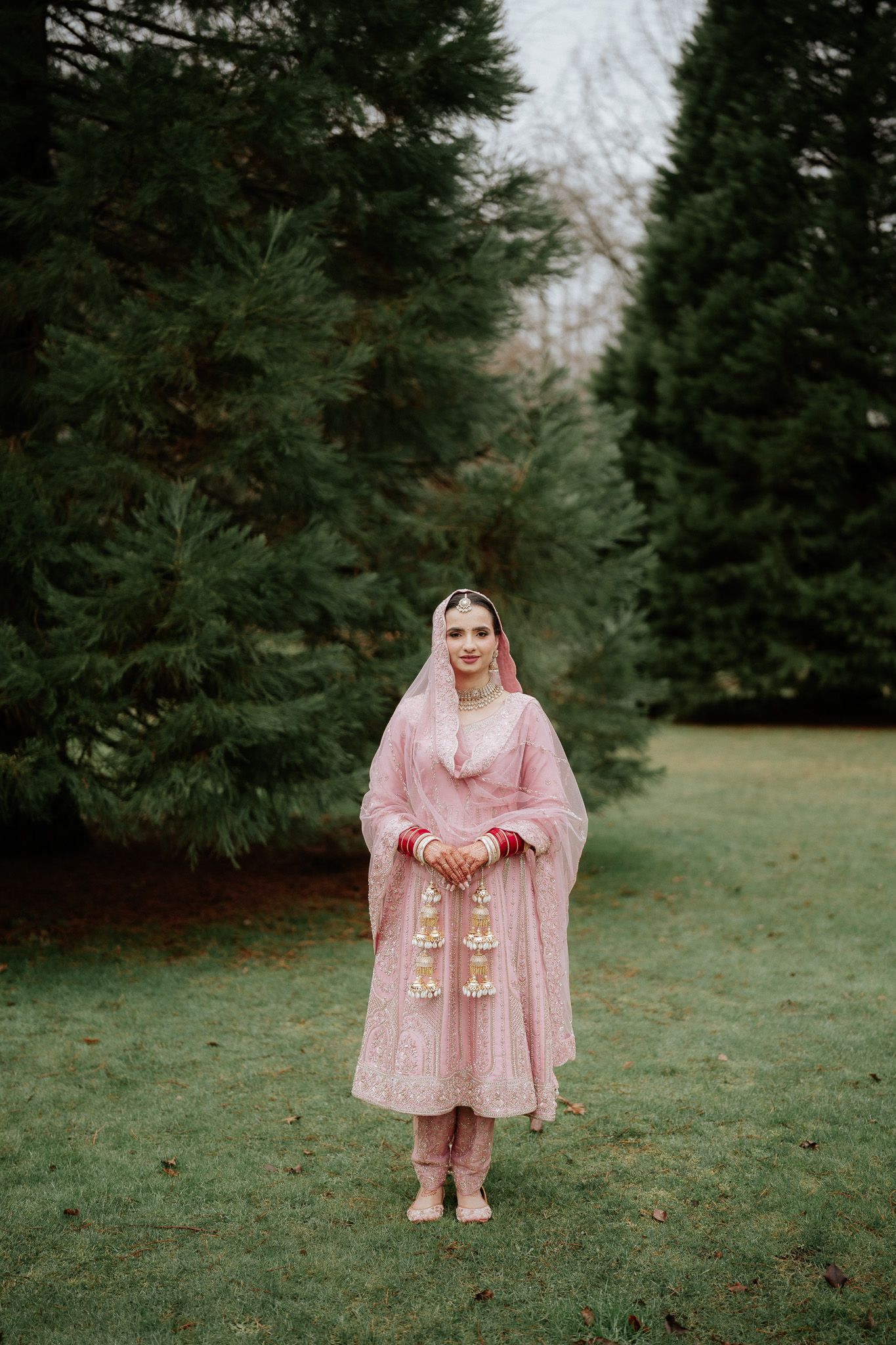 A person in a pink traditional South Asian wedding dress and veil stands between evergreen trees on a grassy lawn.