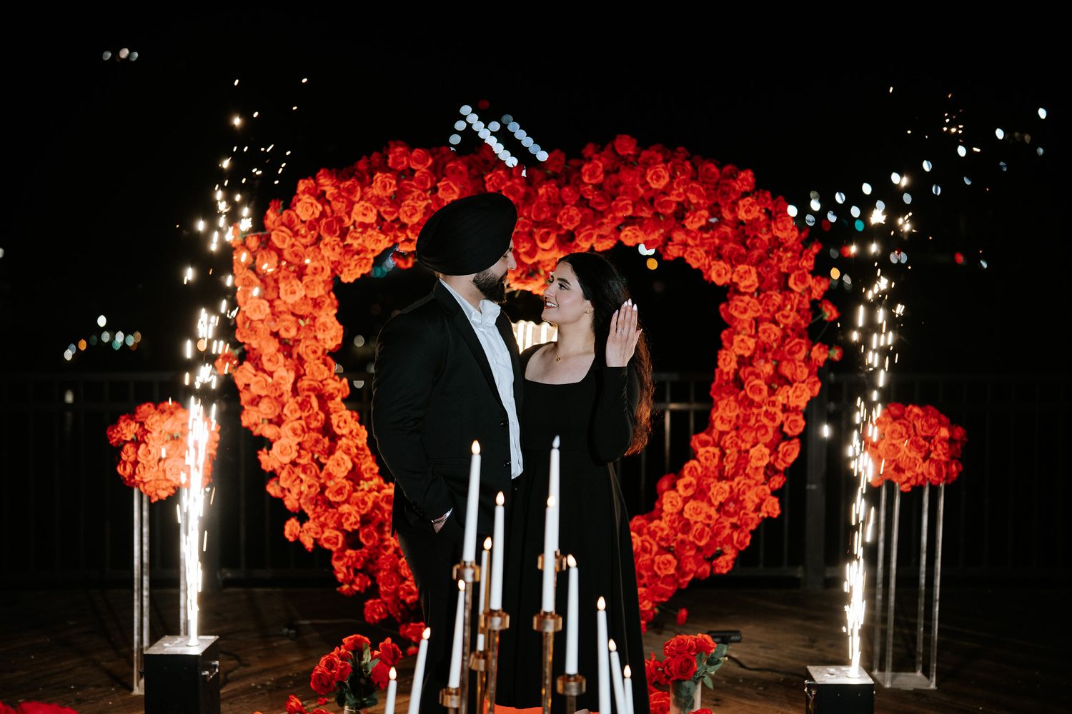A romantic wedding arch made of red roses illuminates a dark outdoor setting with a silhouetted couple standing beneath it.