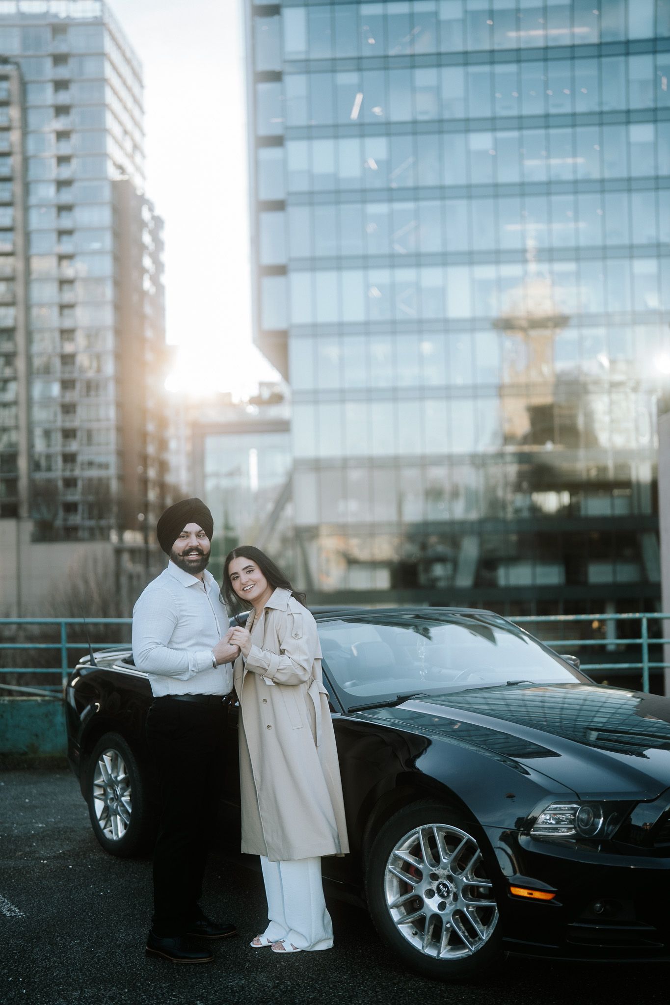 A couple poses beside a sleek black luxury car in front of a modern glass building at sunset.