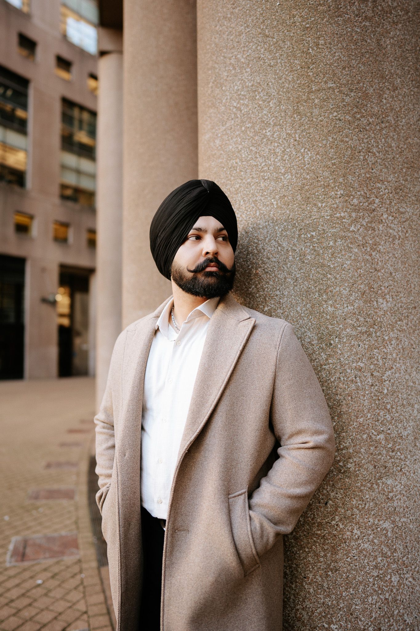 A person wearing a black turban and beige blazer poses against a textured stone column in an urban setting.