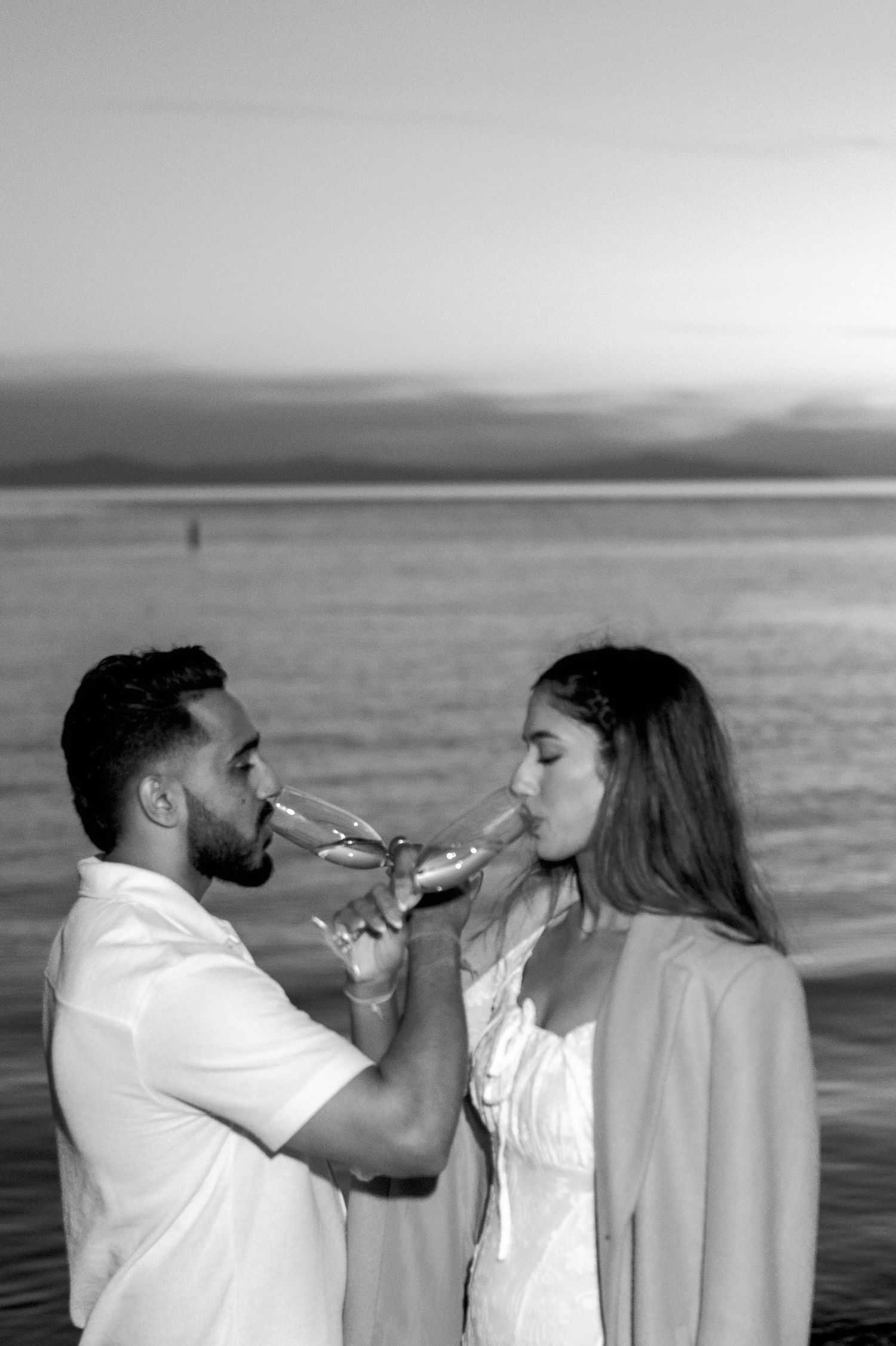 A couple shares a playful moment drinking beverages together on the beach at sunset.