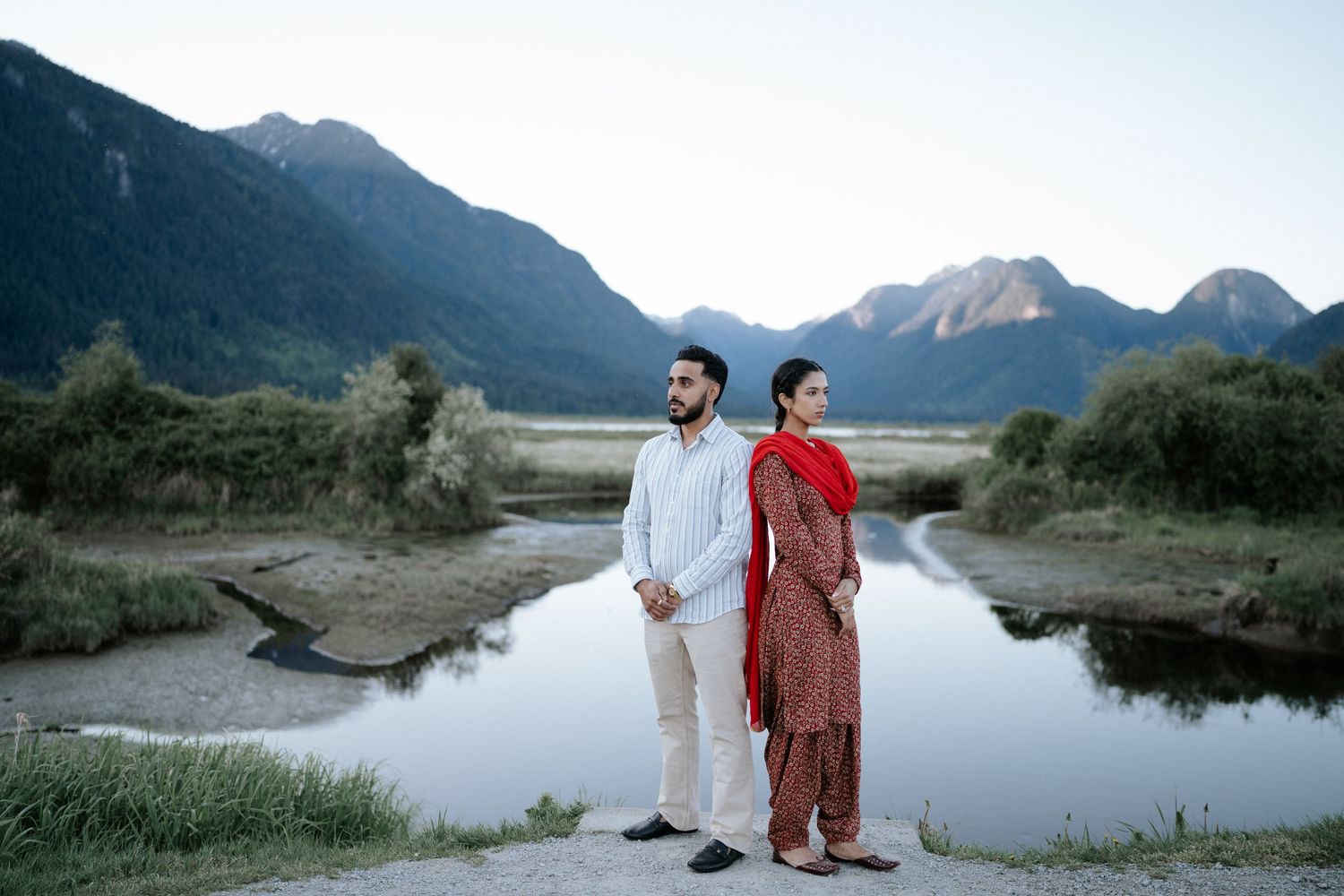 A couple in formal attire poses together in a scenic mountain landscape beside a serene lake at dusk.