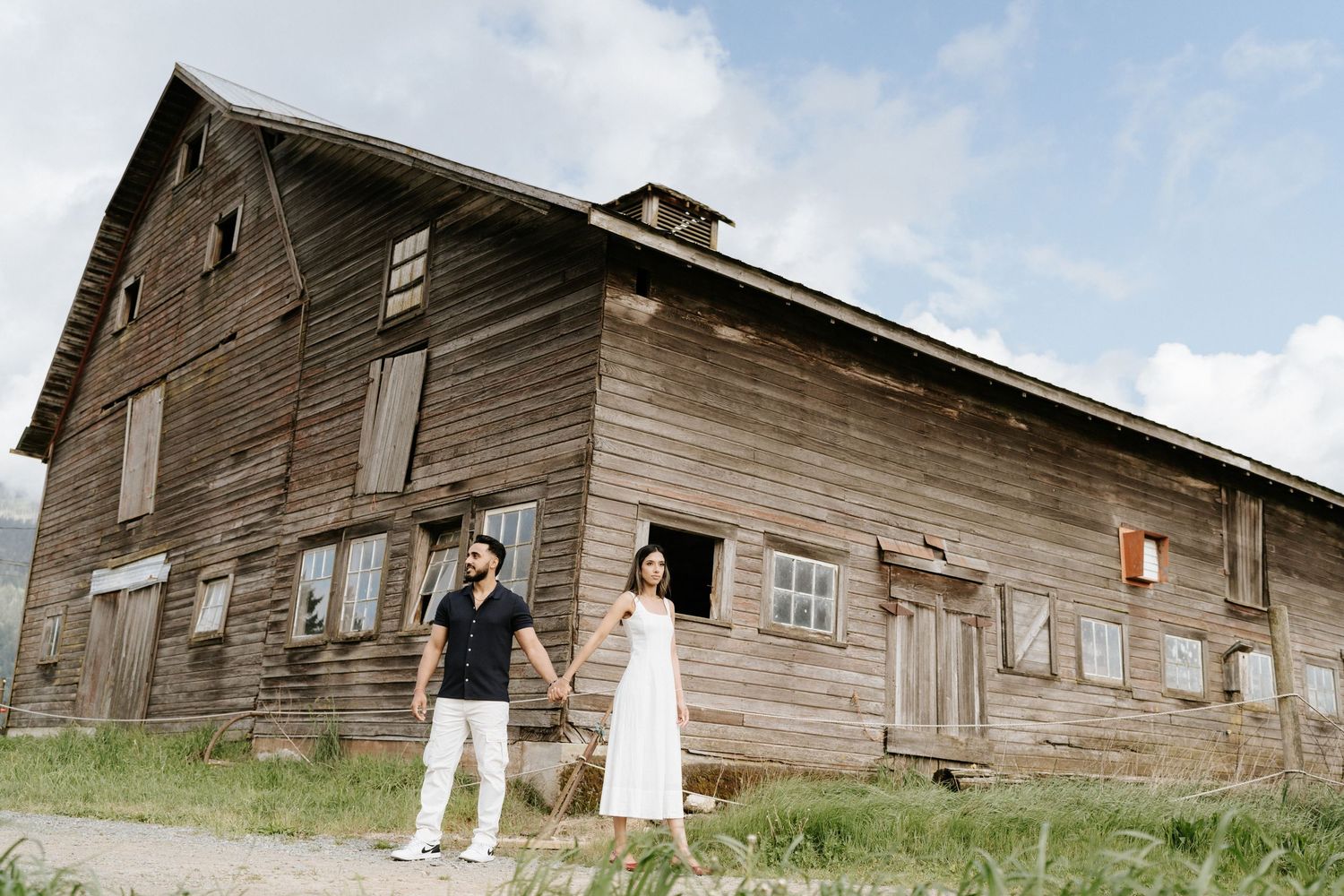 A couple stands in front of a rustic wooden barn with weathered siding on a rural property.