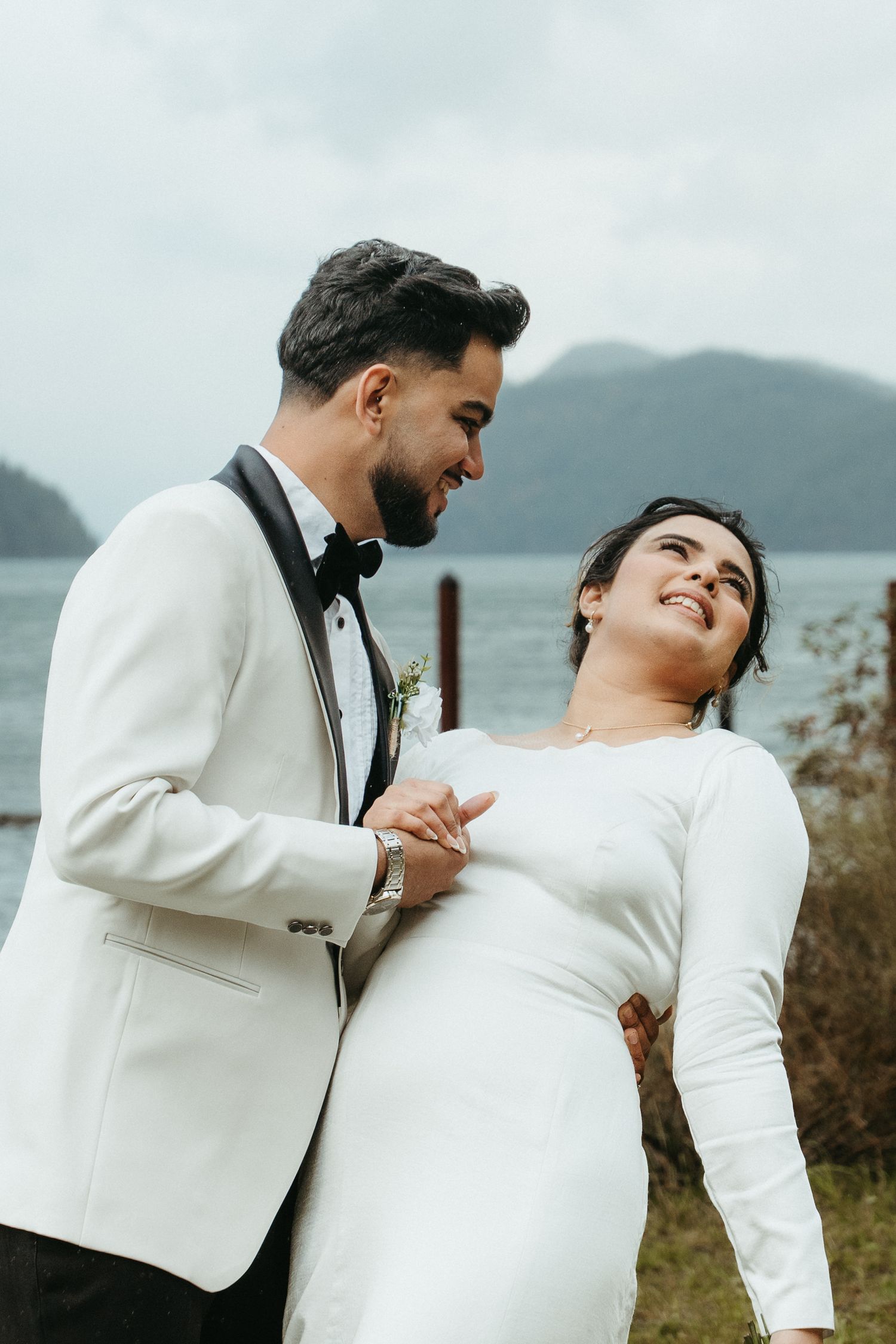 A romantic wedding couple in matching white attire share intimate moments against a scenic mountain backdrop.
