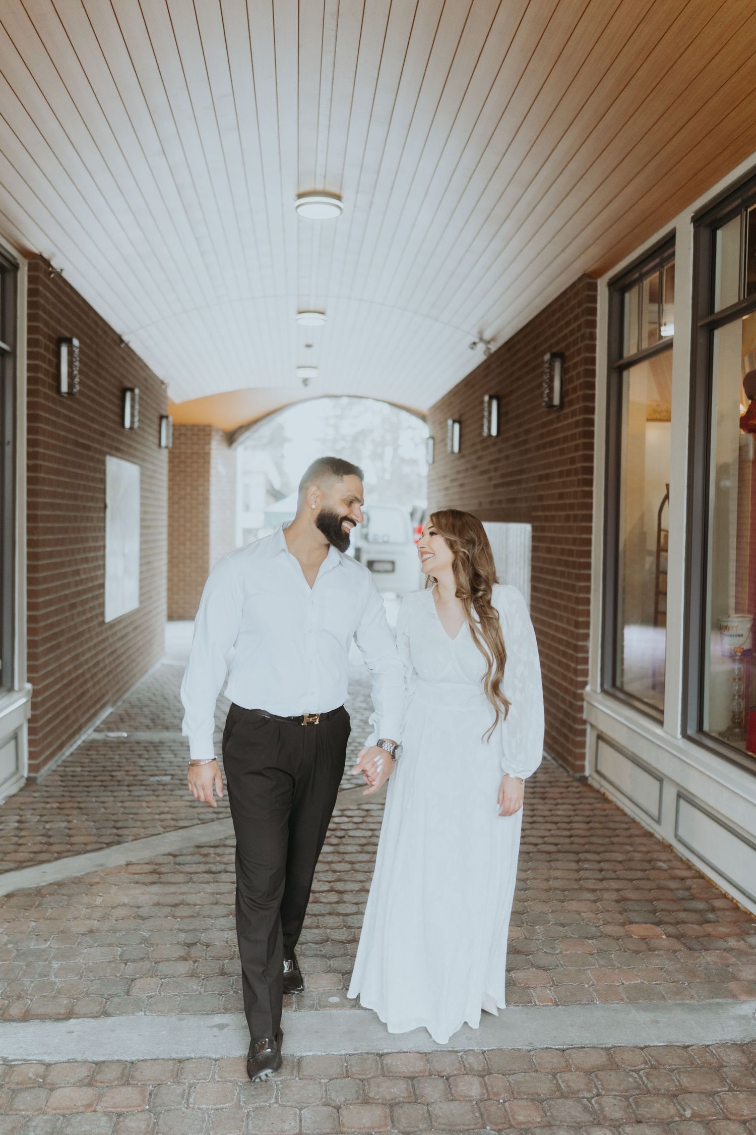 Elegant couple walks through arched brick hallway in contrasting black and white outfits.