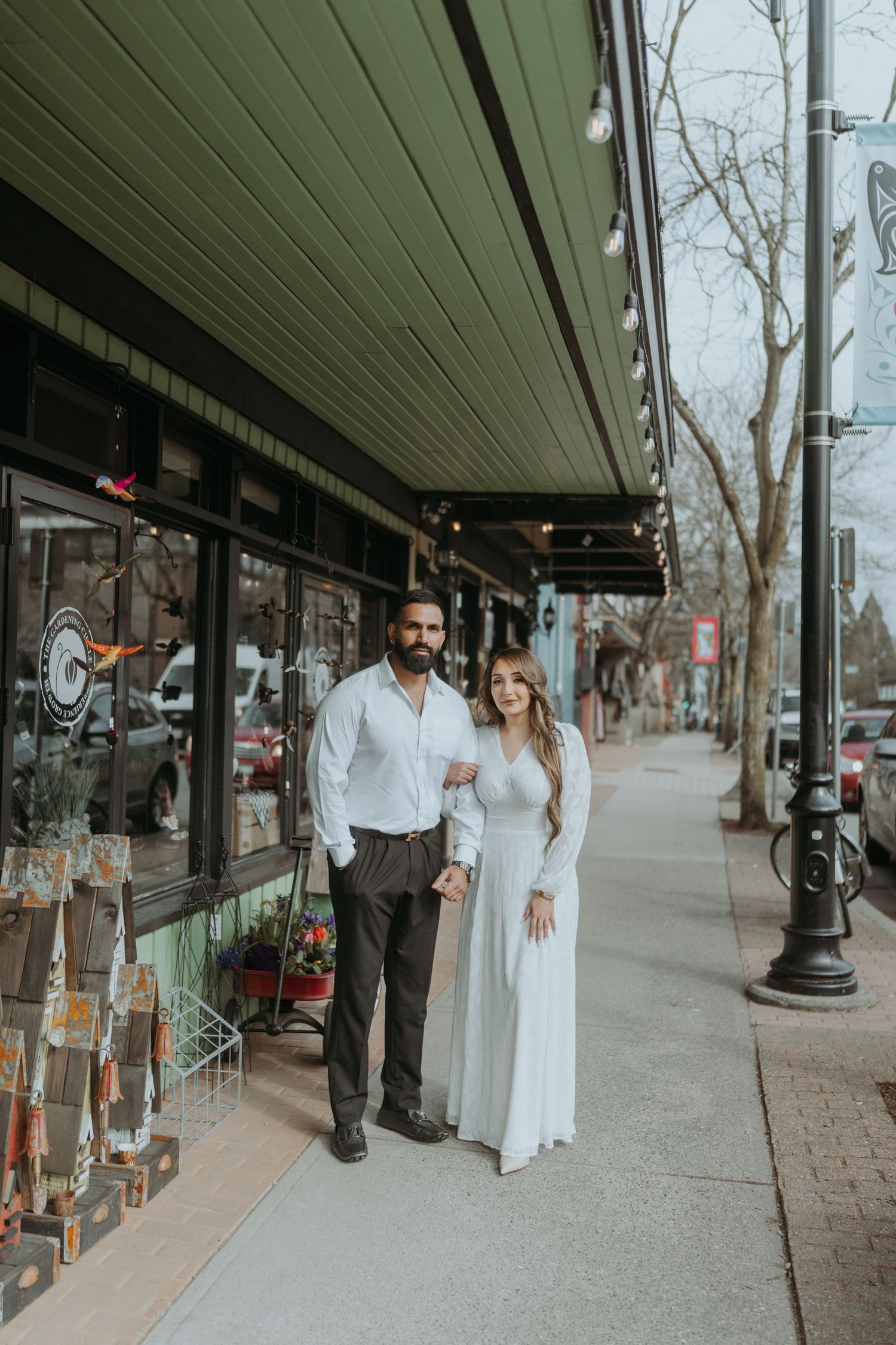 A couple in formal attire poses outside a storefront with green awning and string lights.