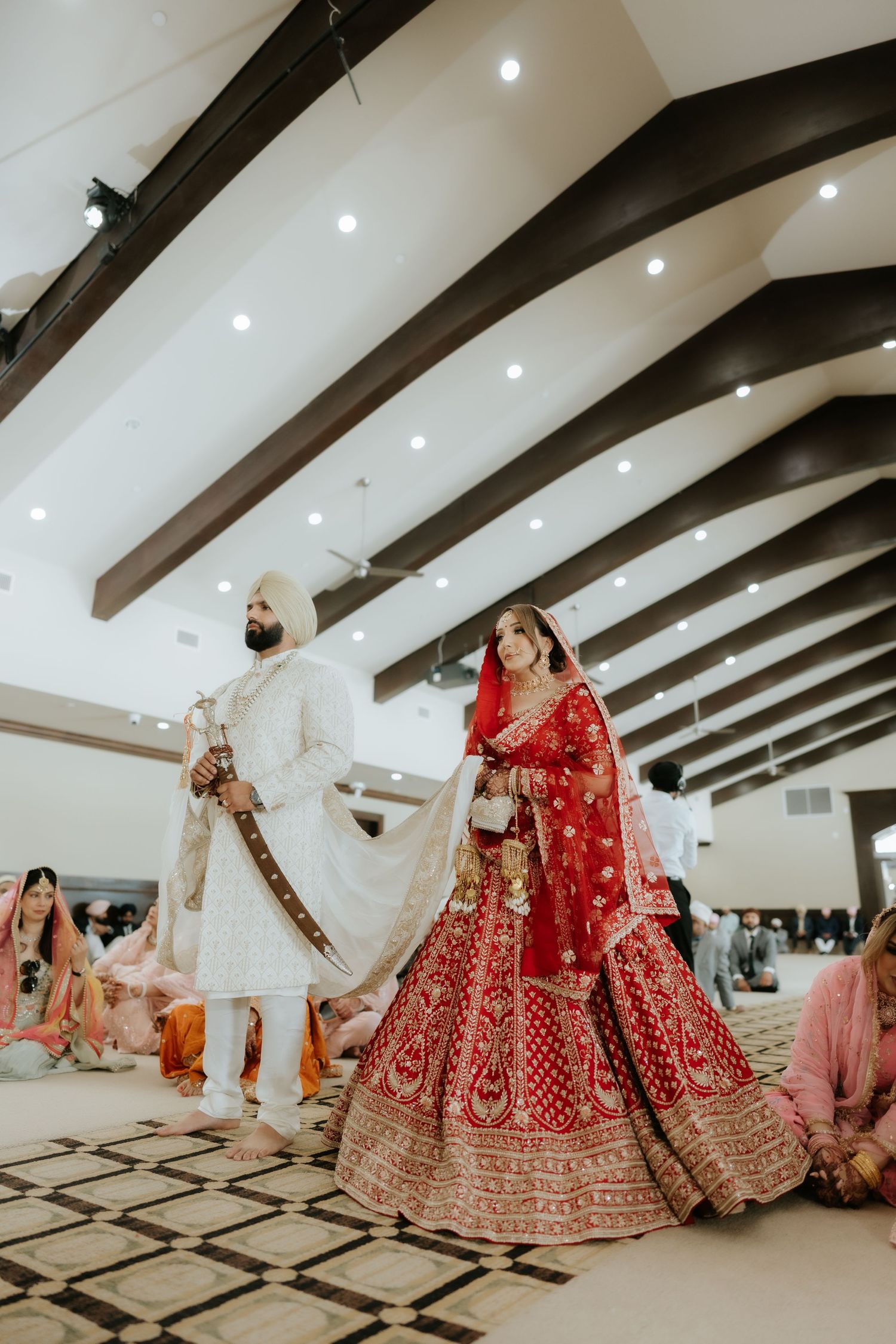 Sikh bride in red lehenga and groom in white sherwani perform wedding ceremony in elegant venue.