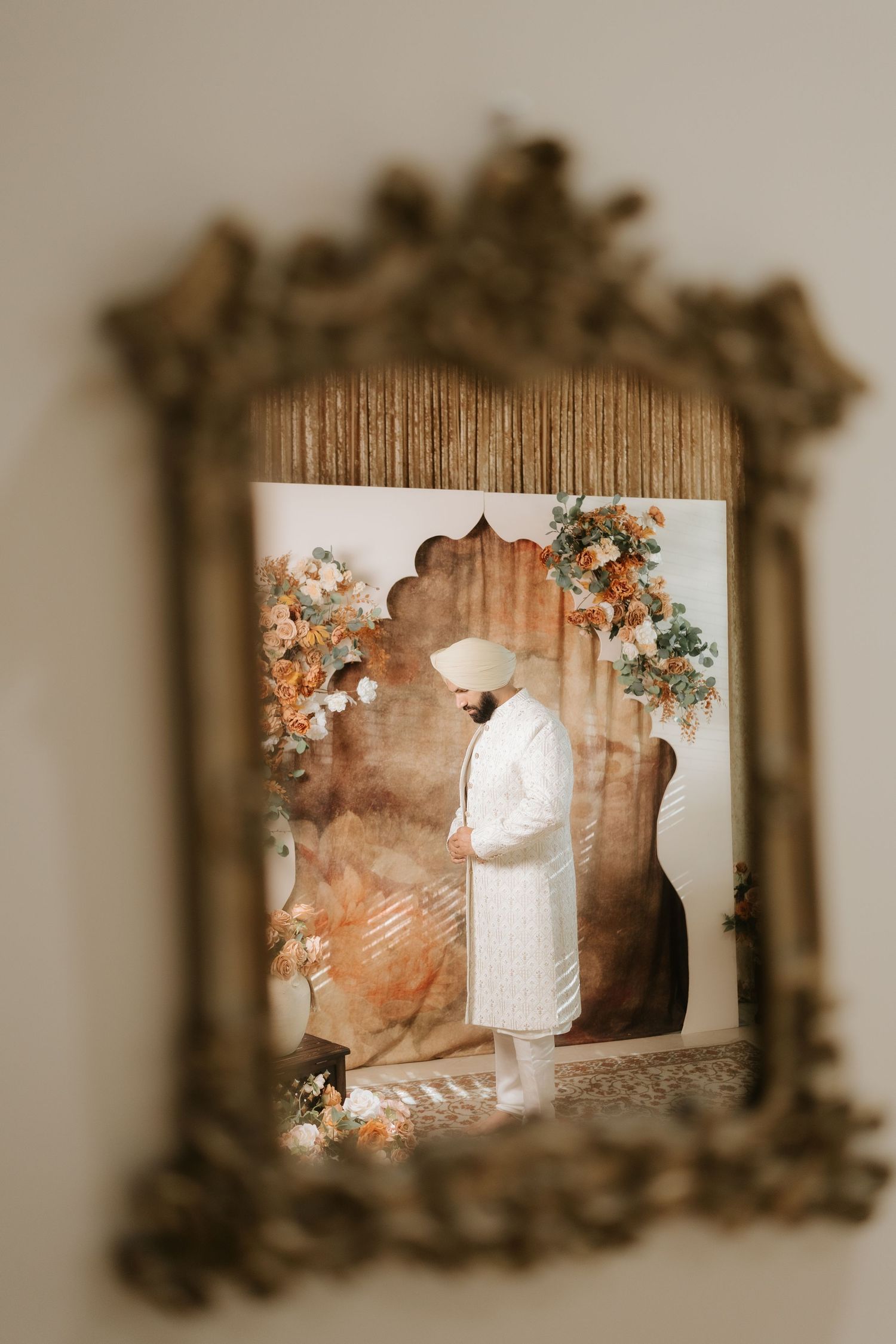 A Sikh groom in white sherwani and turban reflected in an ornate gold mirror with floral backdrop.
