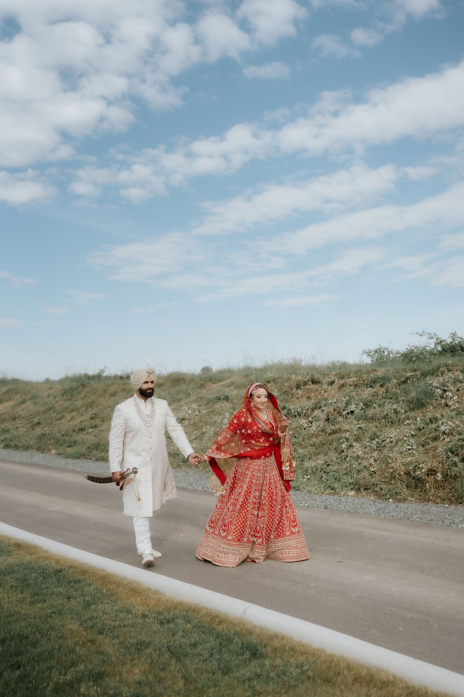 Indian bride in red lehenga and groom in white sherwani walk hand-in-hand on a road under a blue sky.