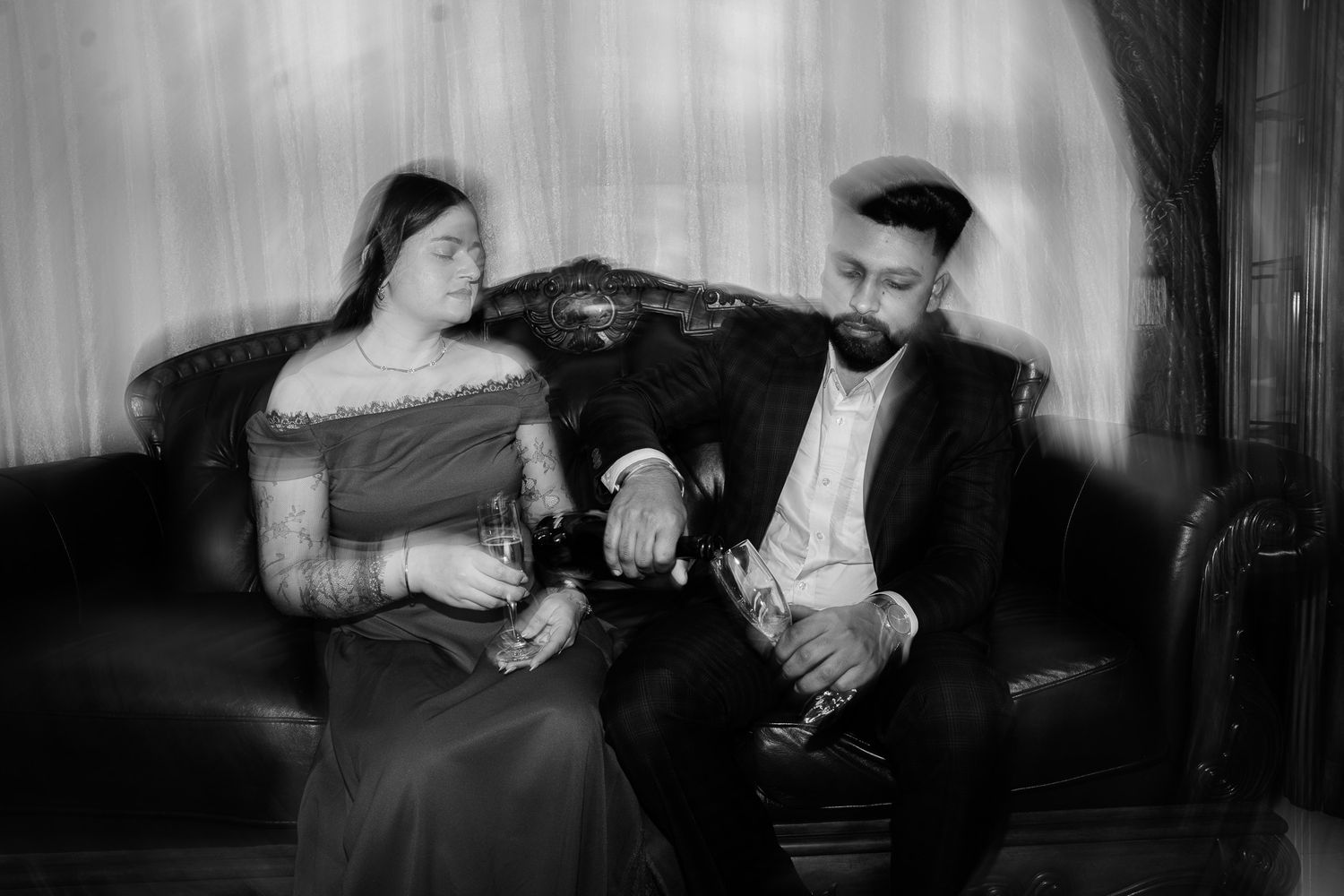 Black and white artistic photo of couple seated on ornate sofa, woman in saree and man in suit with champagne.