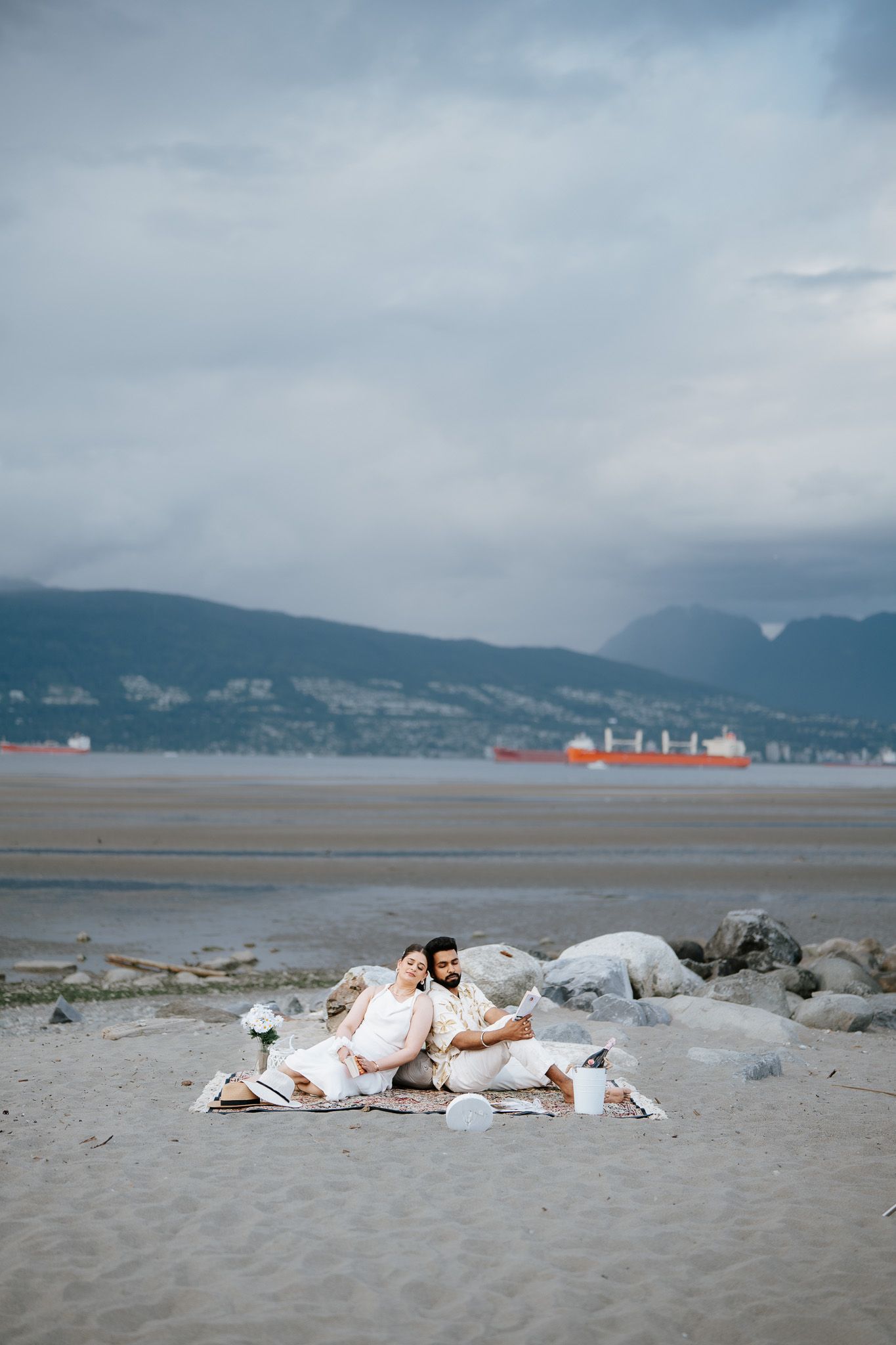 Couple in white outfits sitting on beach blanket with rocky shoreline, mountains, and cargo ship in background.