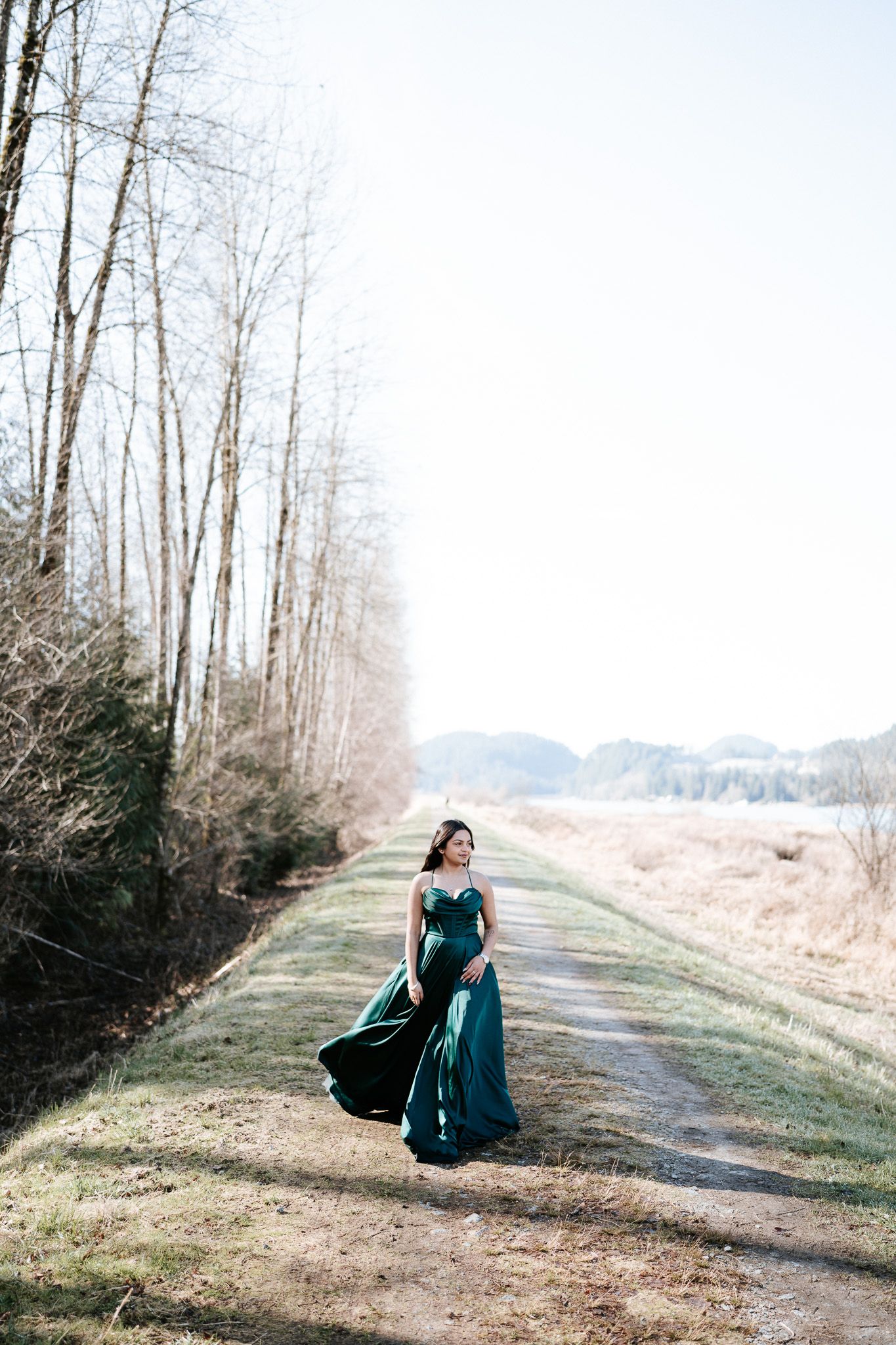 A person in a flowing teal dress walks along a grassy path bordered by bare winter trees on a misty morning.