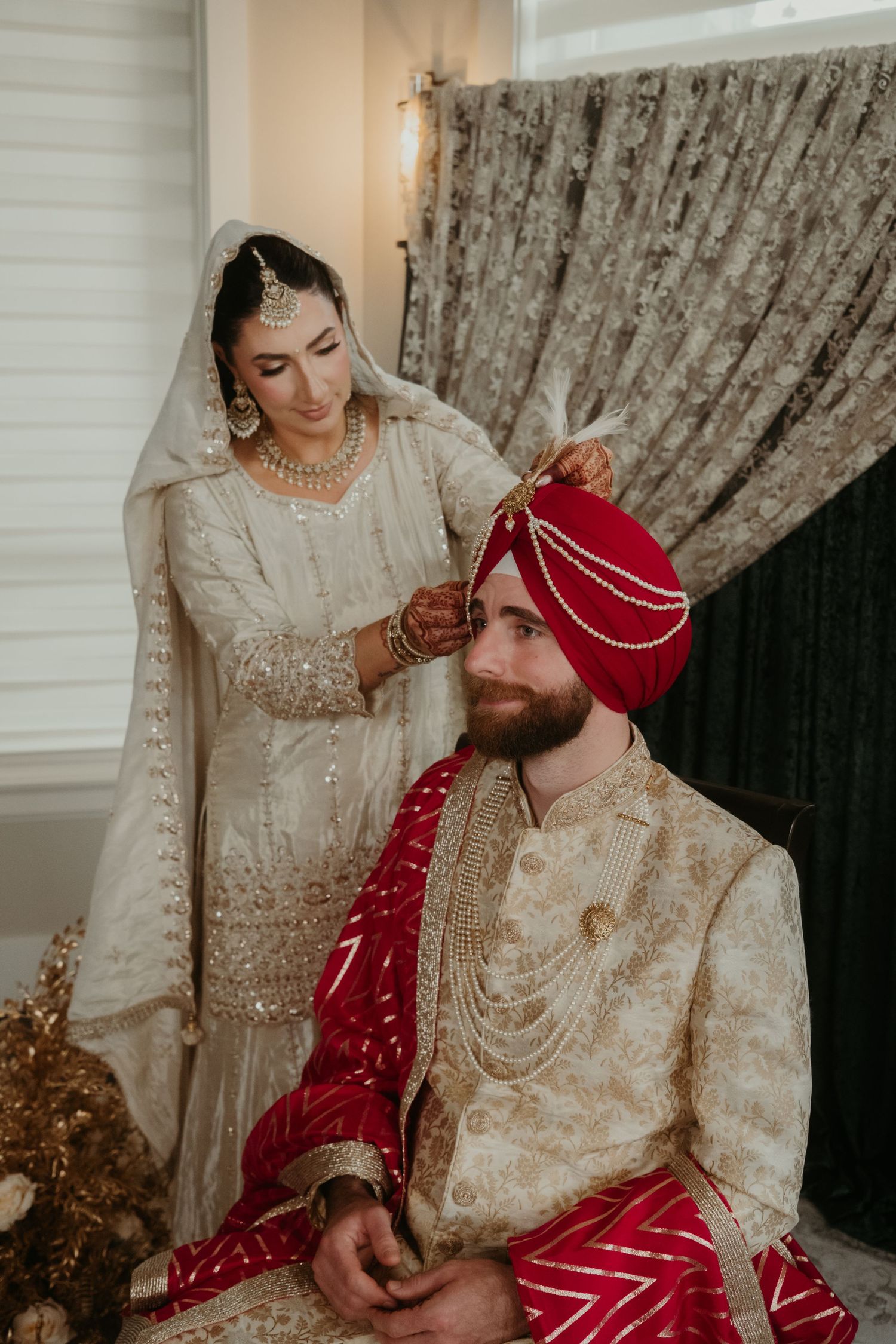 Bride in white lehenga adjusts groom's red turban during traditional South Asian Sikh wedding ceremony.