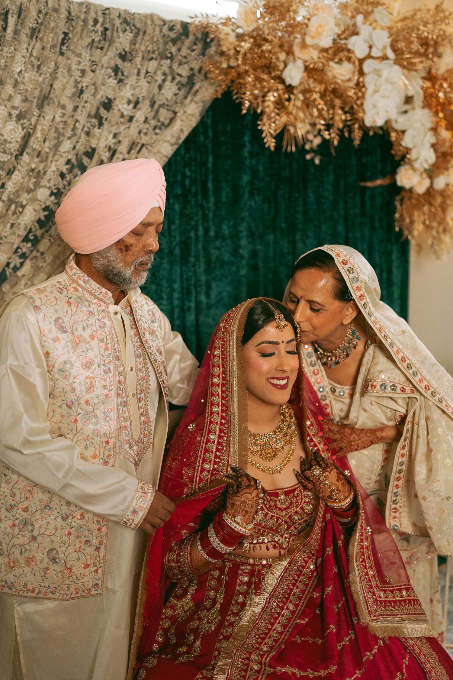 Sikh bride in red lehenga with gold jewelry receives blessings from parents during traditional Indian wedding ceremony.