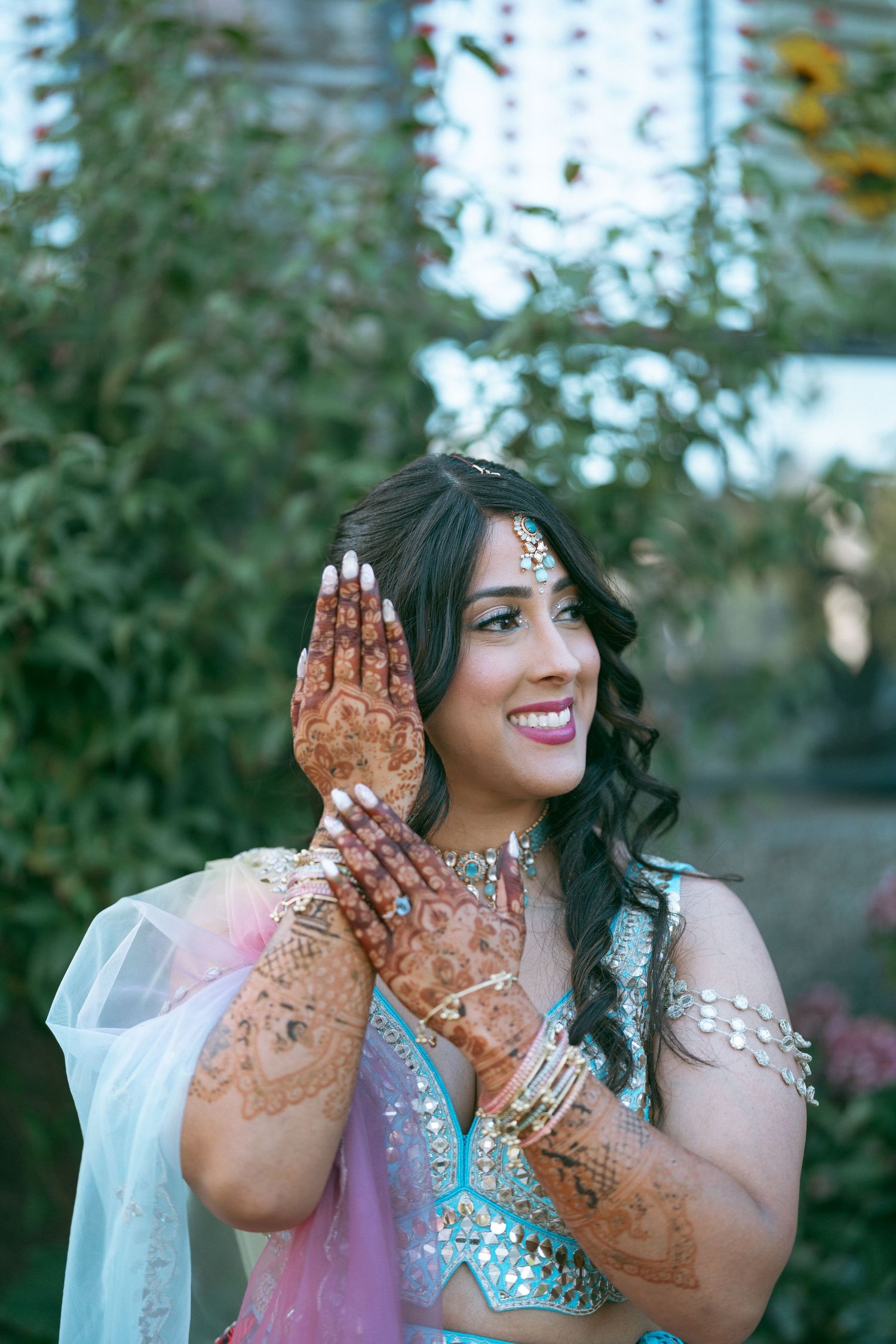 Smiling South Asian bride in turquoise lehenga with intricate henna mehndi designs on hands, wearing traditional jewelry outdoors.
