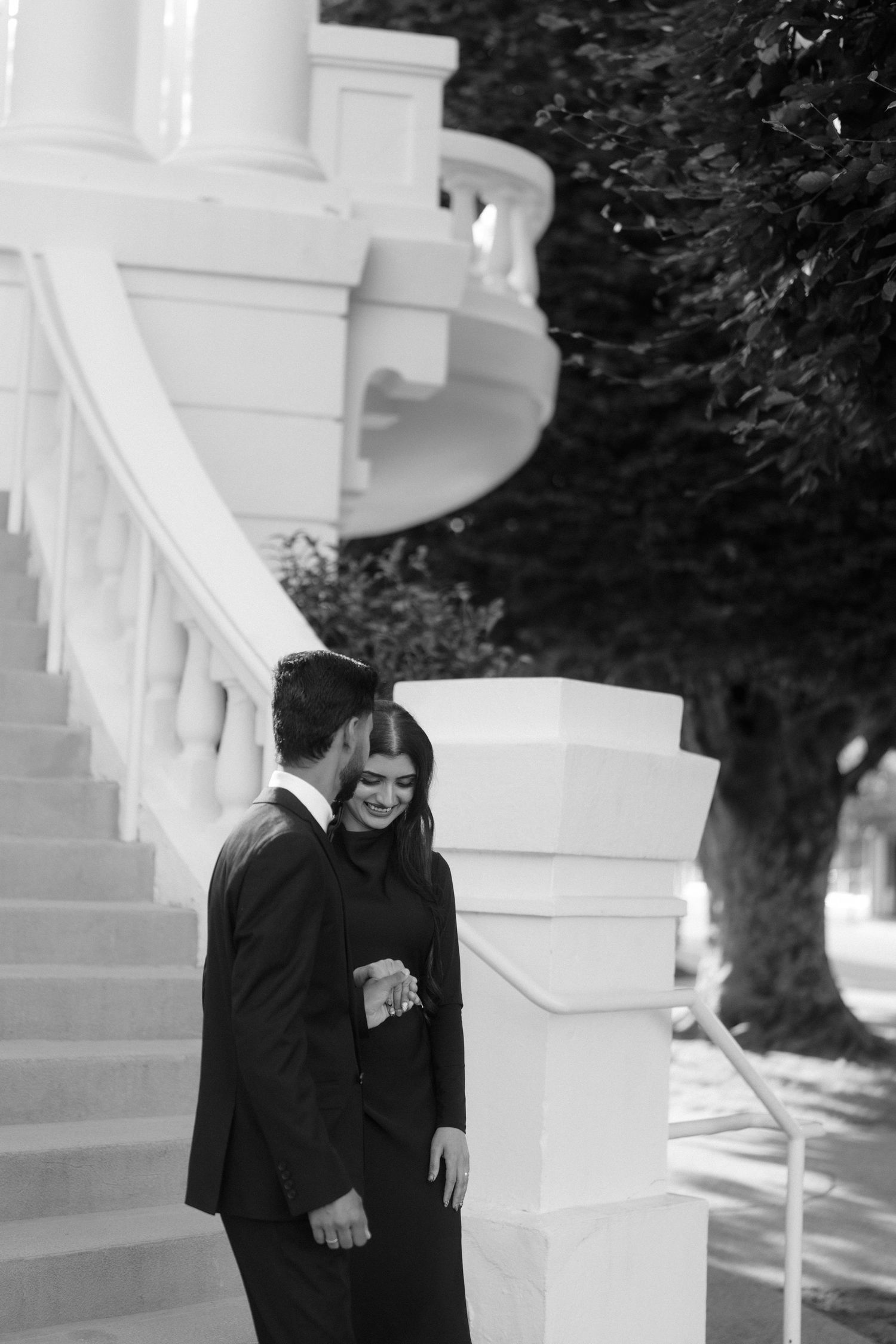 Elegant couple in matching black suits share a romantic moment on grand white staircase in black and white photo.