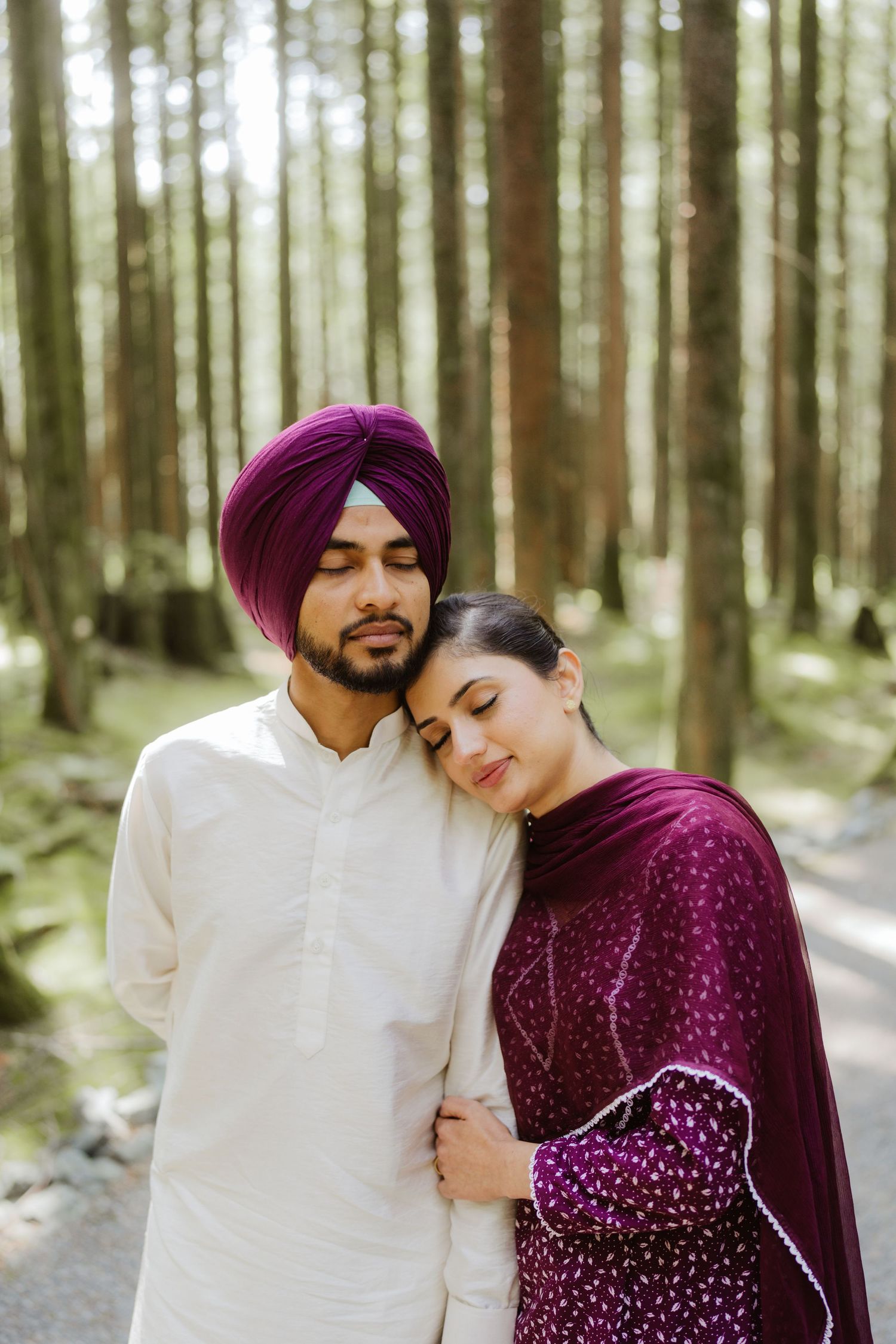 Sikh couple posing in pine forest, man in white kurta and purple turban, woman in matching purple salwar suit.