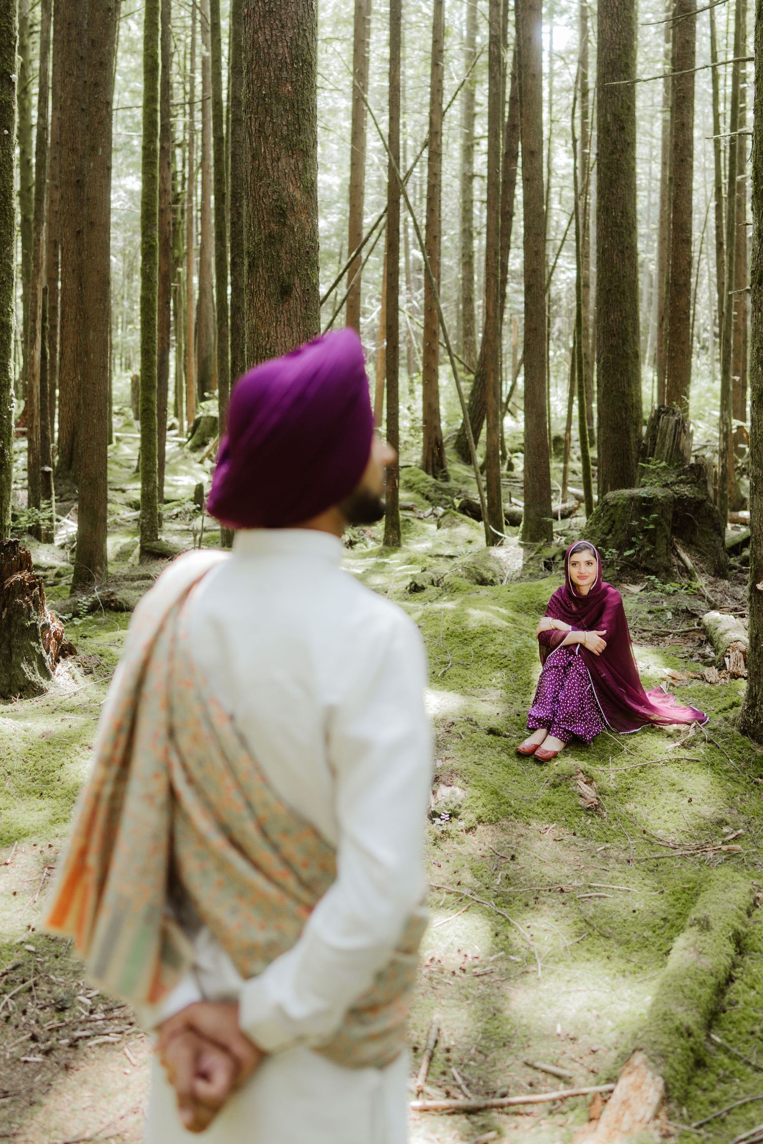 Sikh man in purple turban standing before woman seated on mossy forest floor surrounded by tall pine trees.