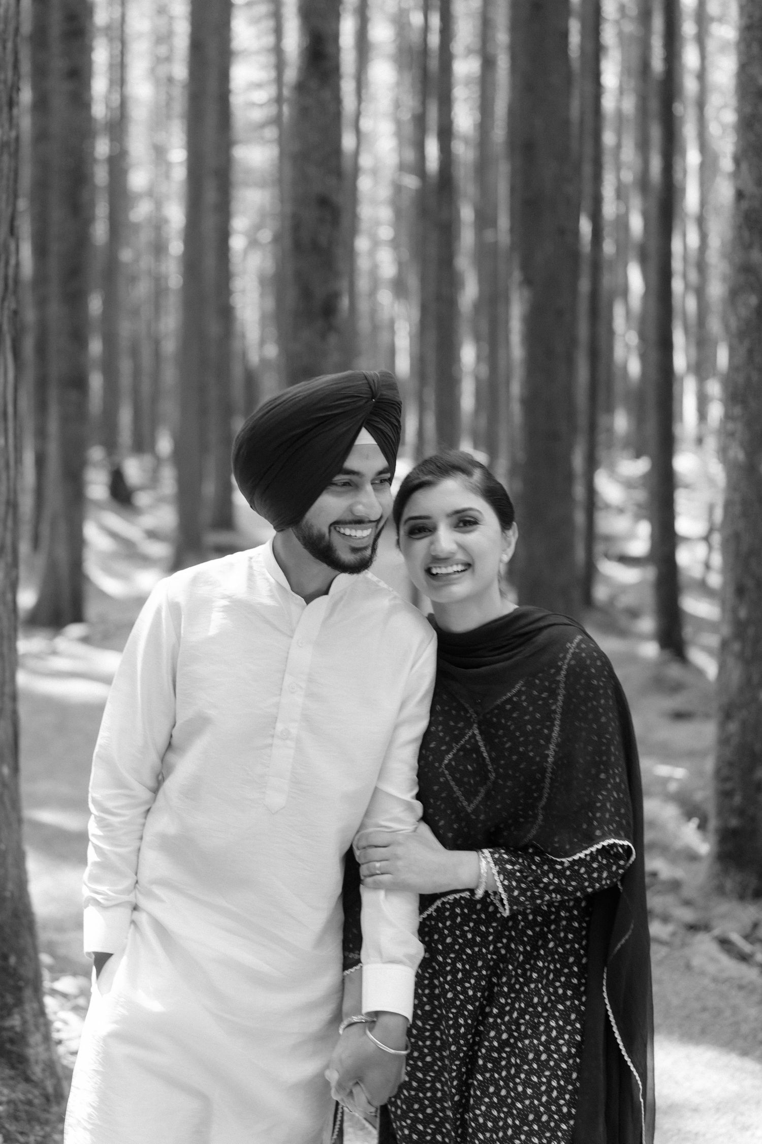 Black and white photo of a smiling Sikh couple in traditional attire posing together in a pine forest.