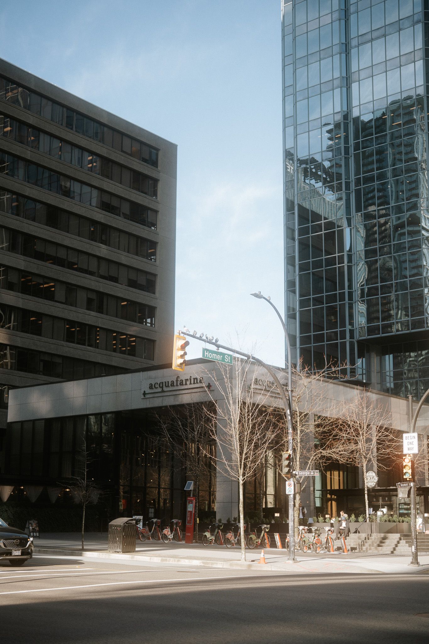 Modern glass office buildings line an urban street corner with bare winter trees and reflective facades.