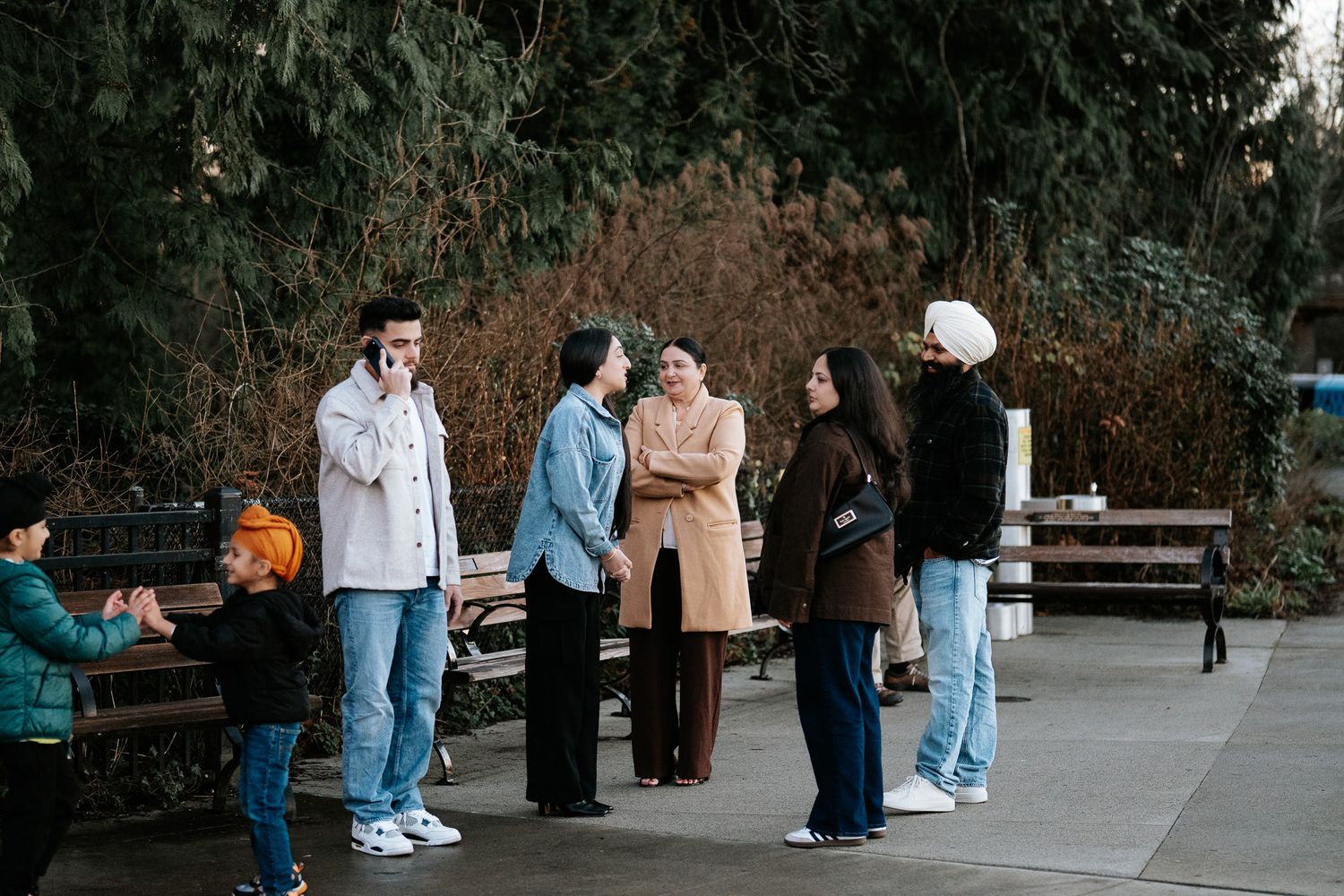 A group of people walking and gathering in a park setting with trees and benches visible in the background.