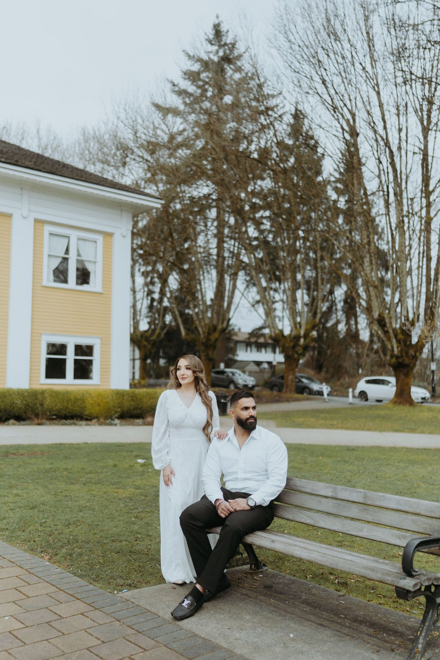 A couple sits together on a park bench with a yellow building and trees in the background.