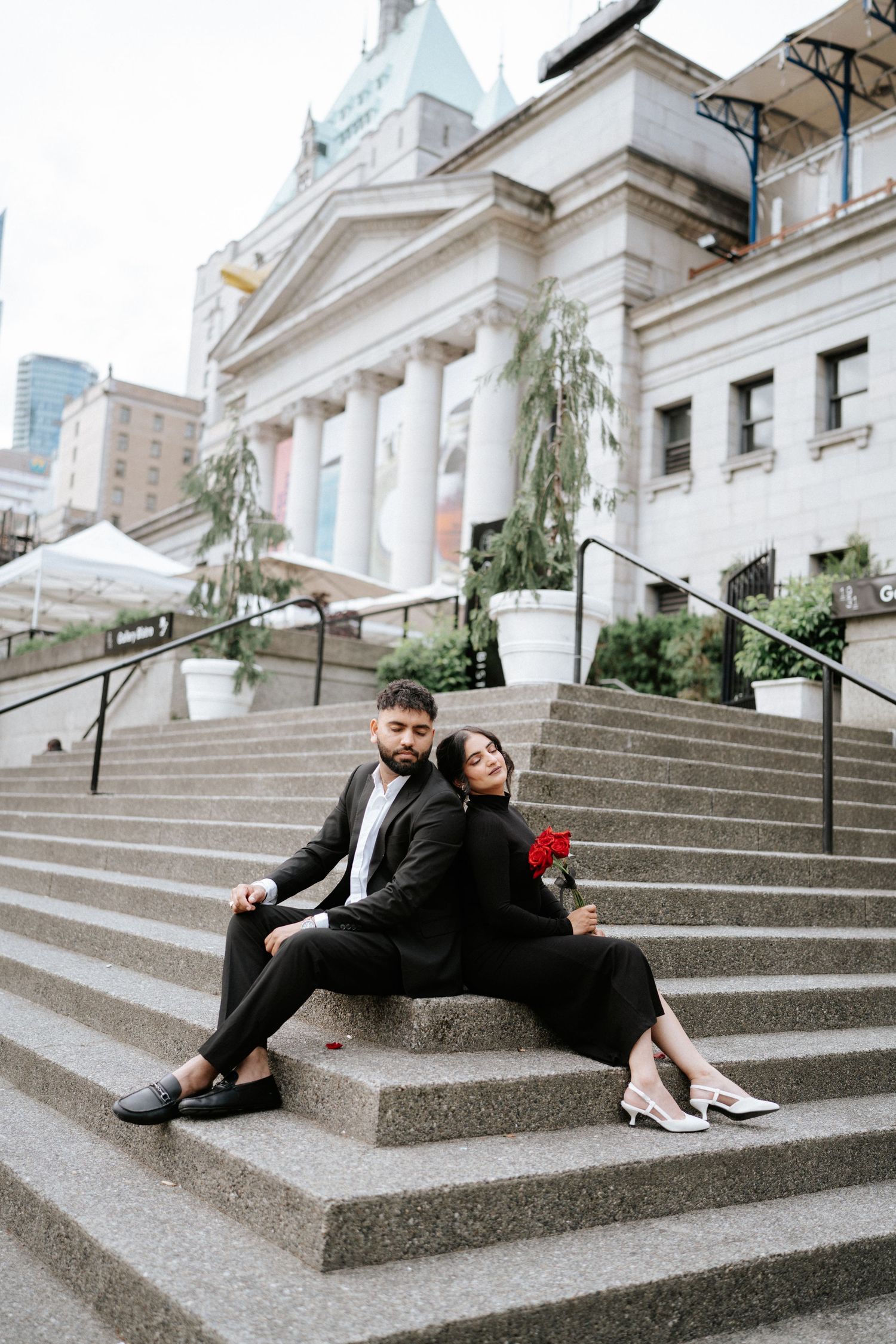 A couple in formal black attire sits romantically on grand stone steps of a historic building.