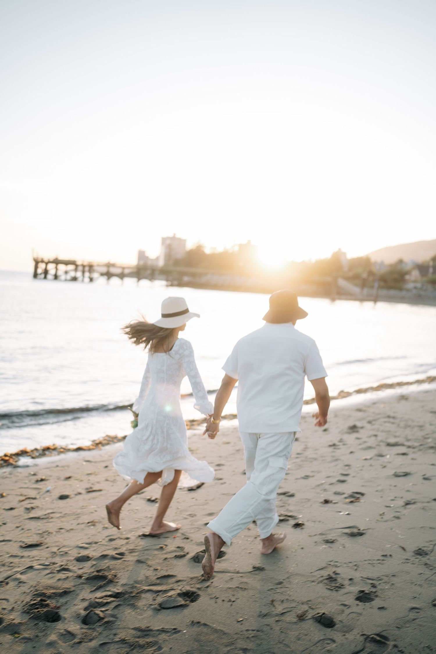 Two people in white outfits run playfully along a beach at sunset, creating a romantic silhouette against the golden light.