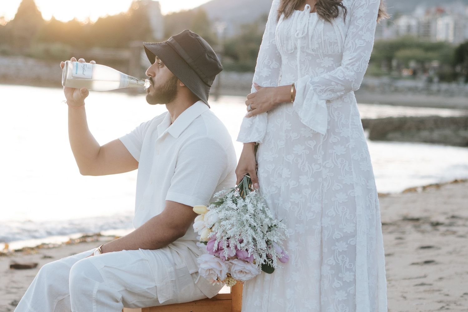 A couple in white wedding attire embraces on a beach at sunset during their romantic coastal ceremony.