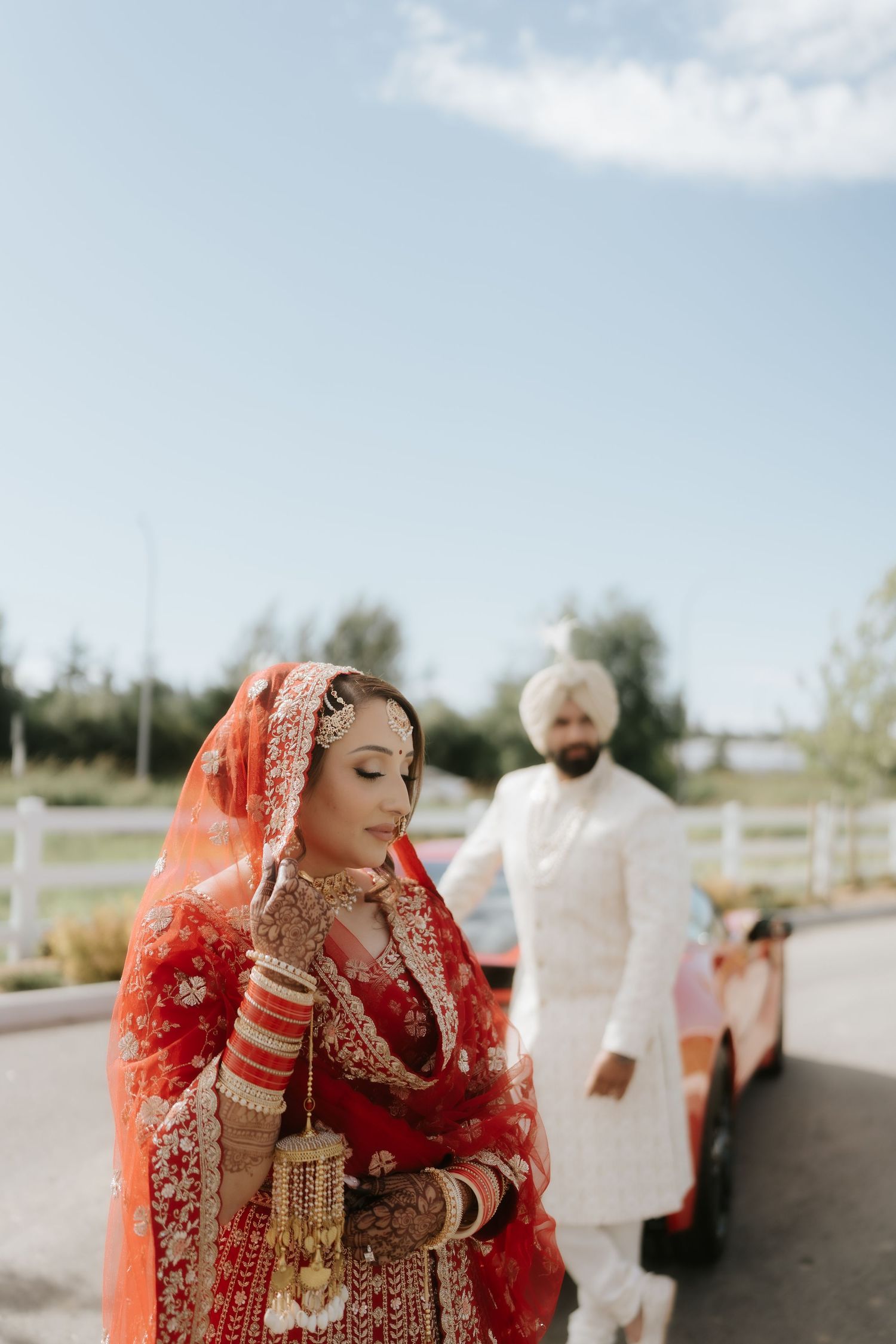 Indian bride in red lehenga with groom in white sherwani and turban posing outdoors on sunny wedding day.