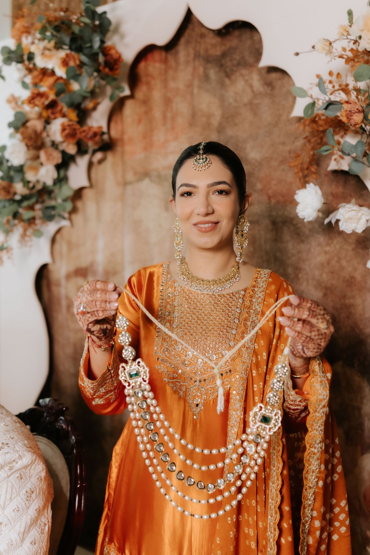 Bride in orange salwar kameez holds layered gold jewelry, wearing maang tikka and earrings at Indian wedding ceremony.