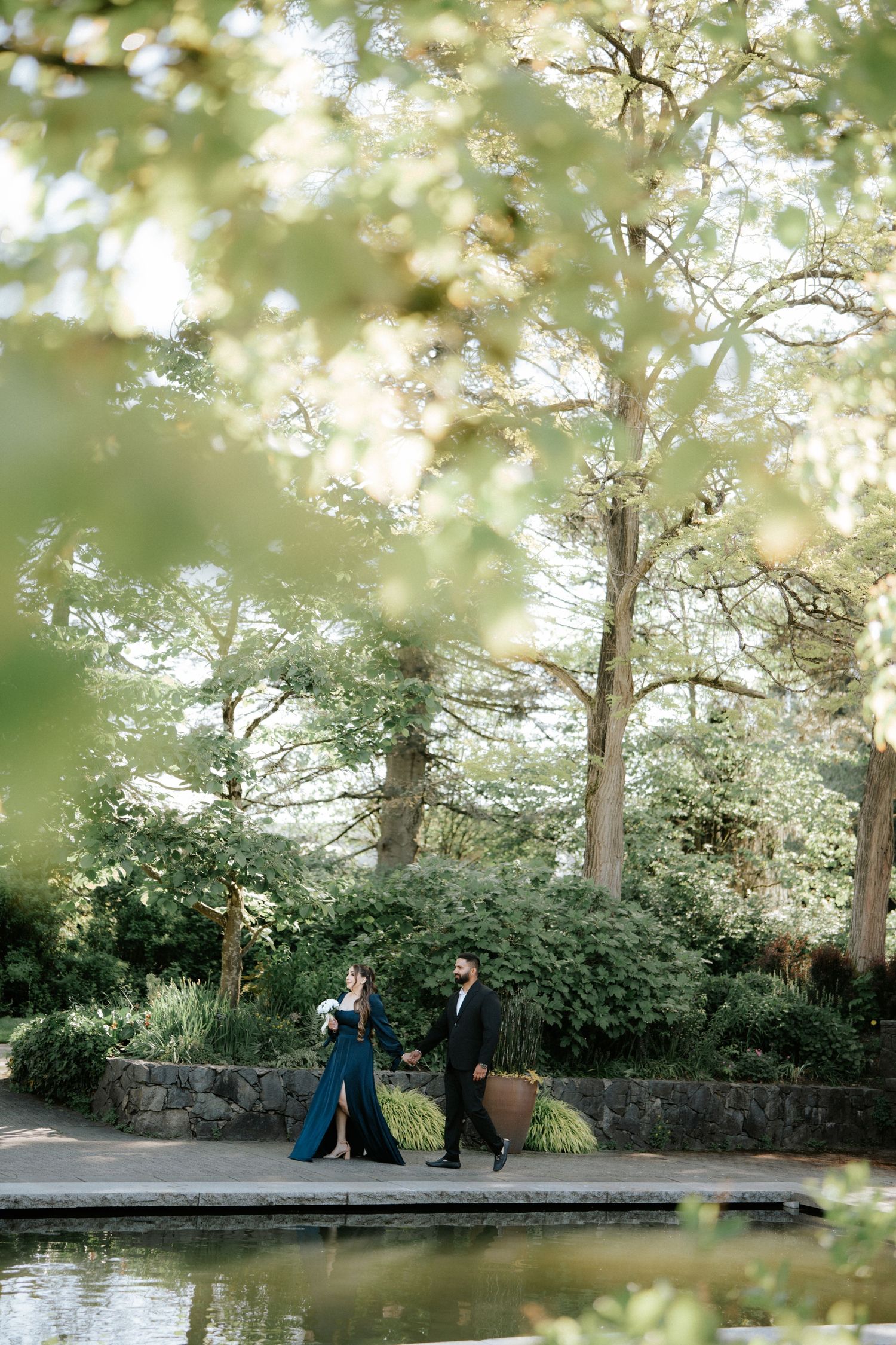 Couple walks hand in hand through lush green garden by a pond, woman in elegant teal gown, man in black suit.