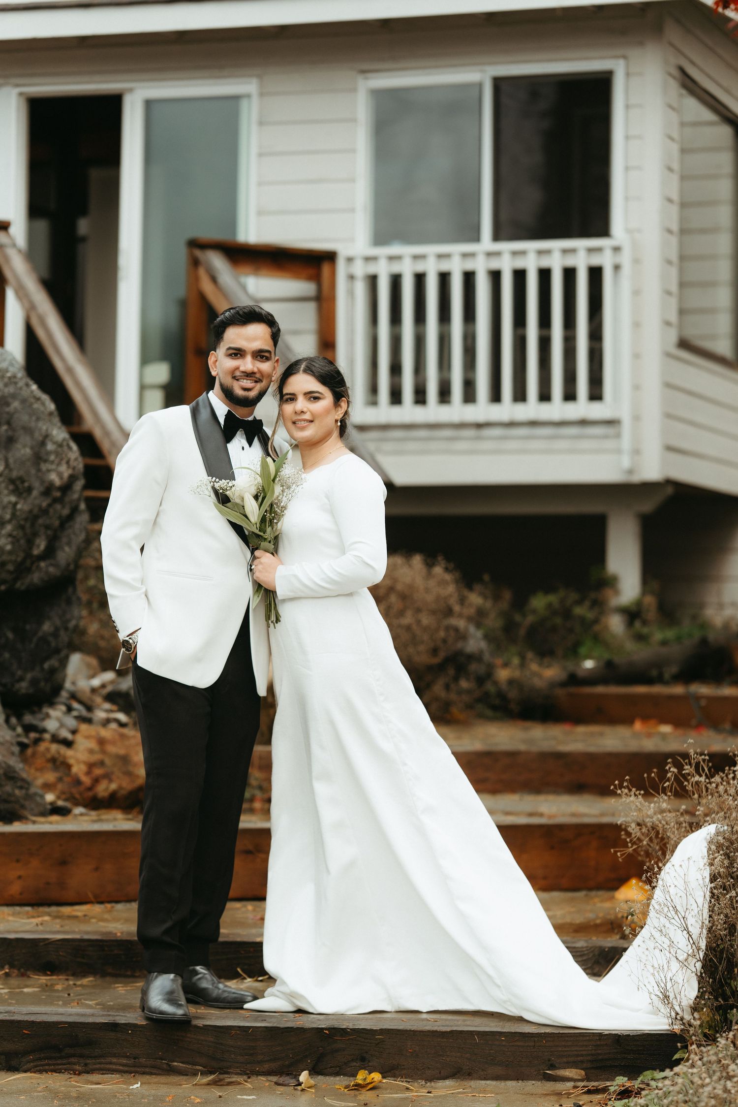 A couple in formal wedding attire poses together in front of a white house on a rainy day.