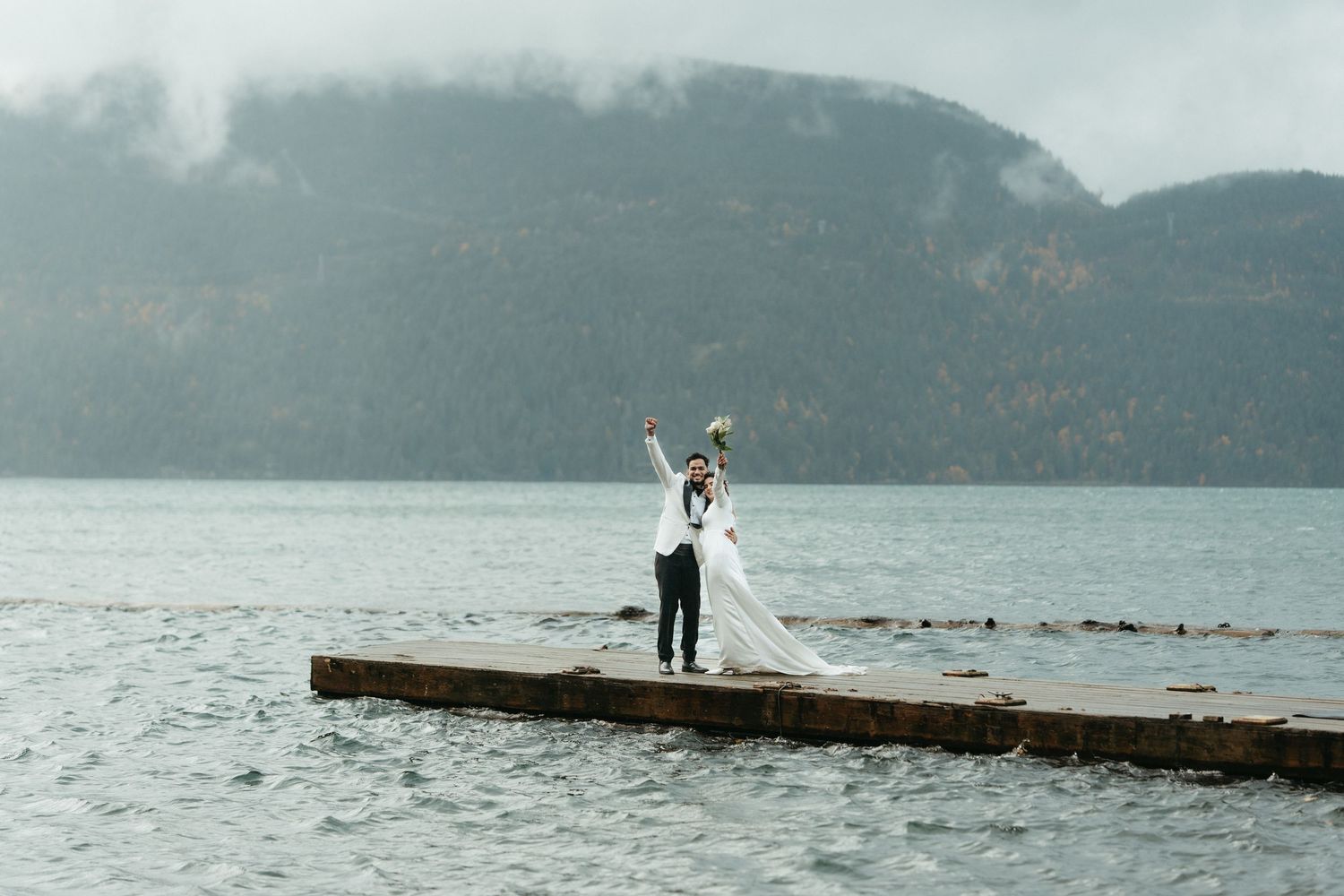 A couple in wedding attire stands on a wooden dock overlooking a scenic mountain lake on their special day.