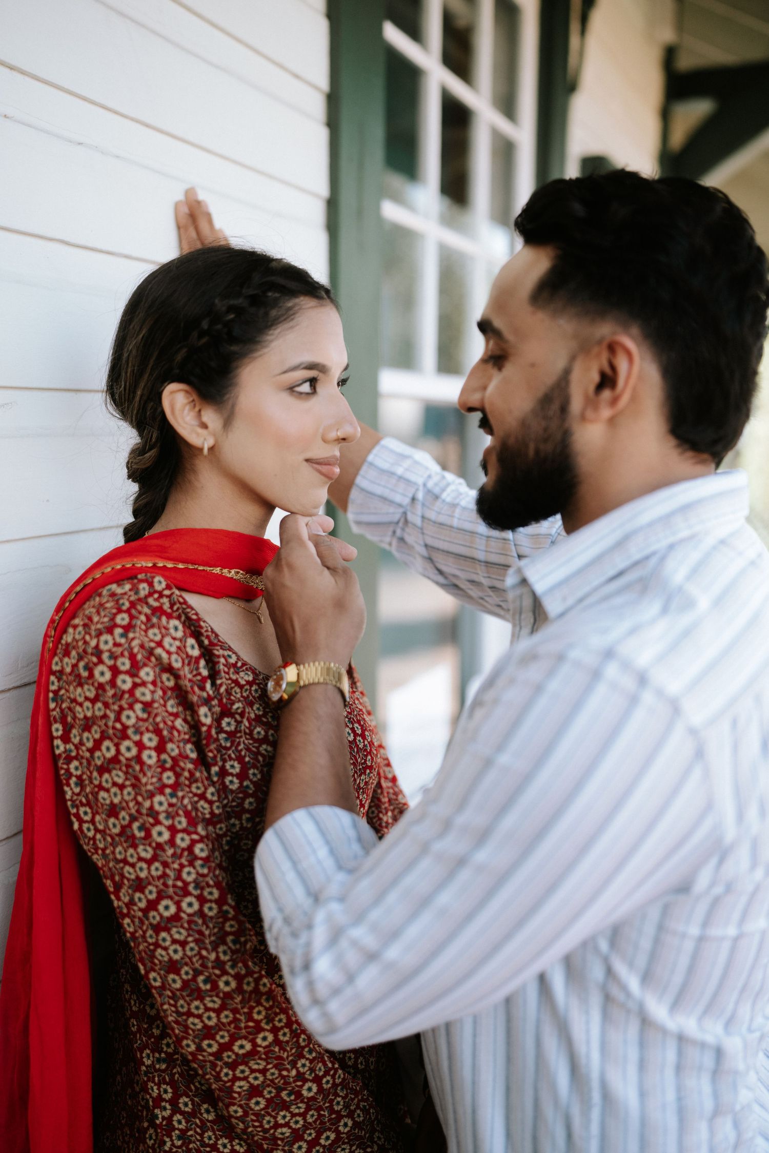 A romantic couple in traditional South Asian attire share an intimate moment against a white wall.