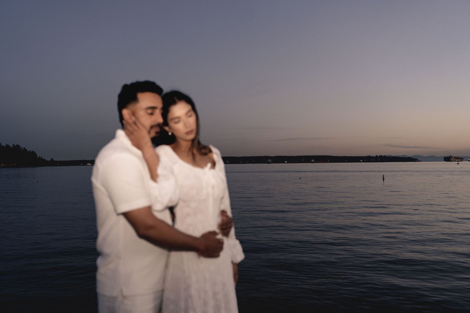 A couple in white clothing embraces by the waterfront at sunset, with a calm ocean and dusky sky in the background.