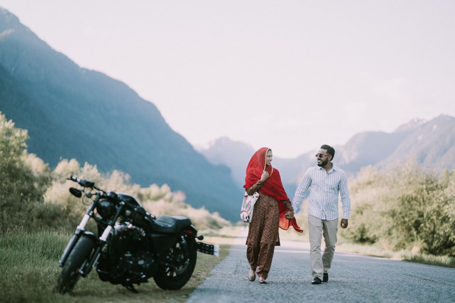 A couple walks hand in hand on a mountain road with a parked motorcycle, set against scenic mountain landscape.
