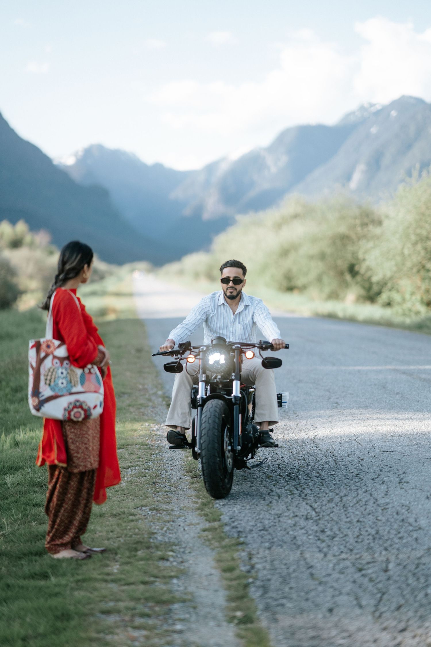 A motorcycle rider and companion in red dress stand on a scenic mountain road surrounded by majestic peaks.