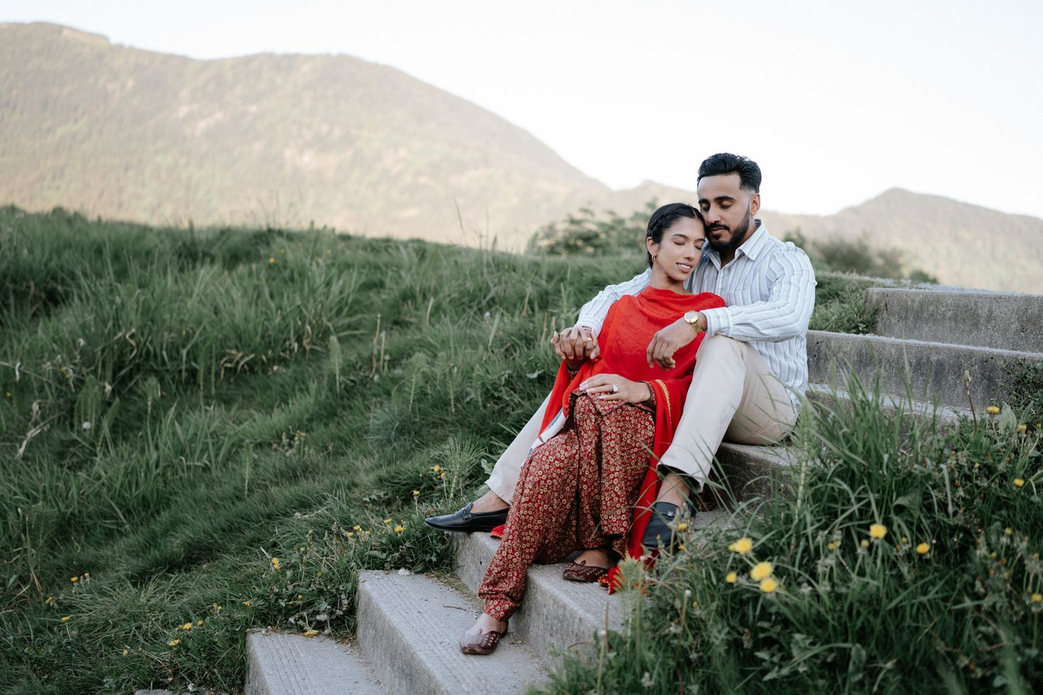 Couple in traditional Indian attire sharing romantic moments during outdoor photoshoot in grassy hillside setting.