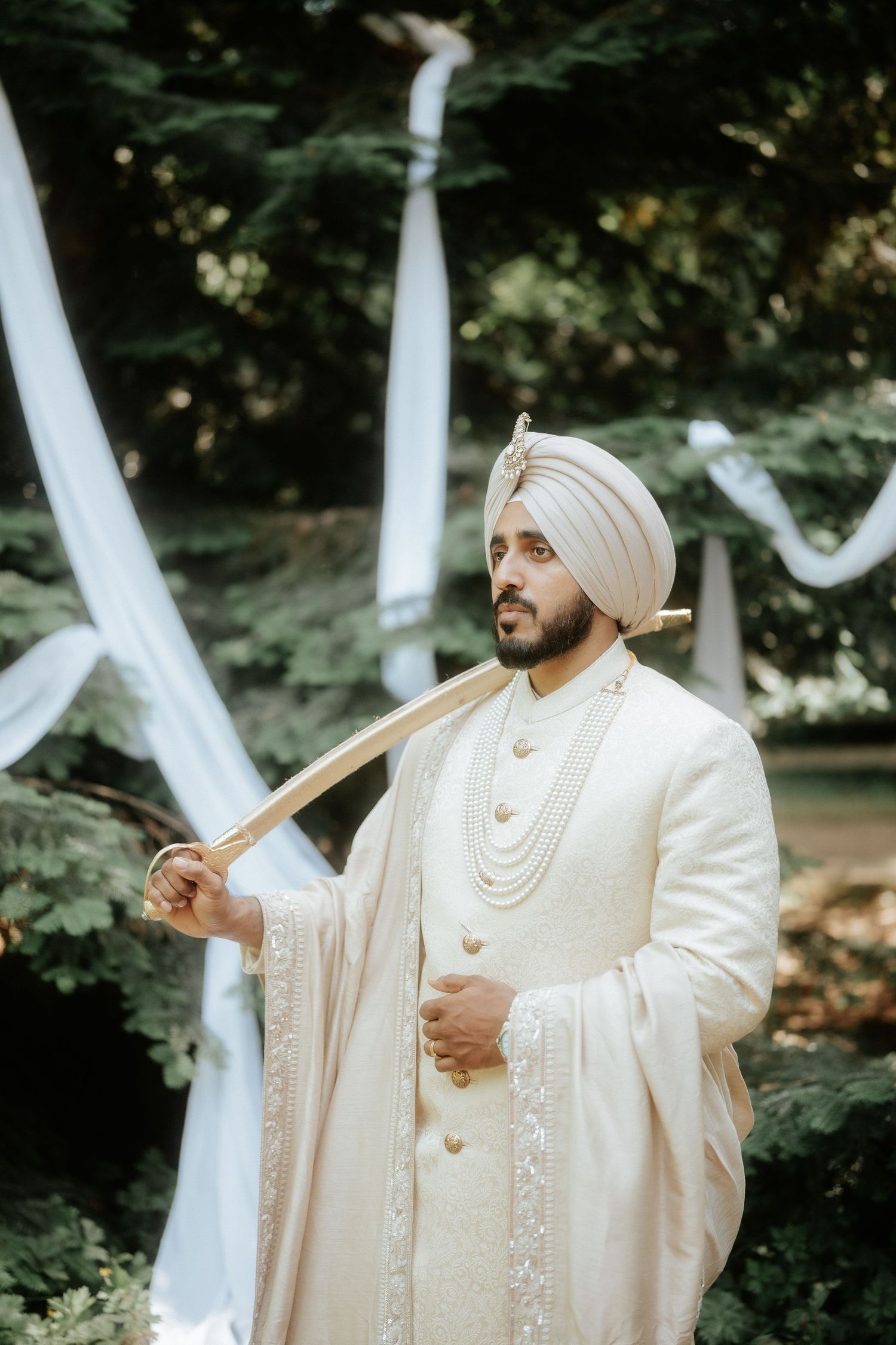 A groom in cream-colored traditional Sikh wedding attire poses against draped white fabric outdoors.
