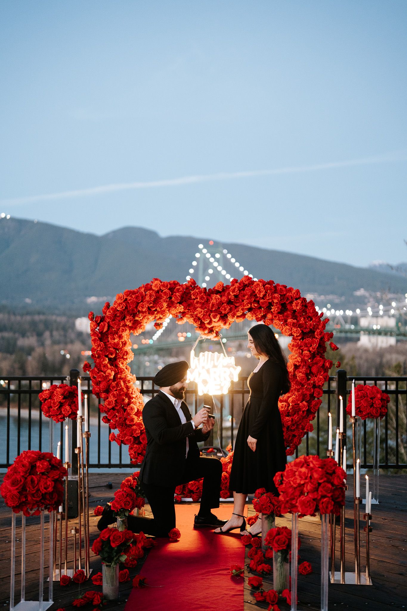 Red floral arches illuminate a romantic waterfront proposal setting at sunset with mountains in the background.